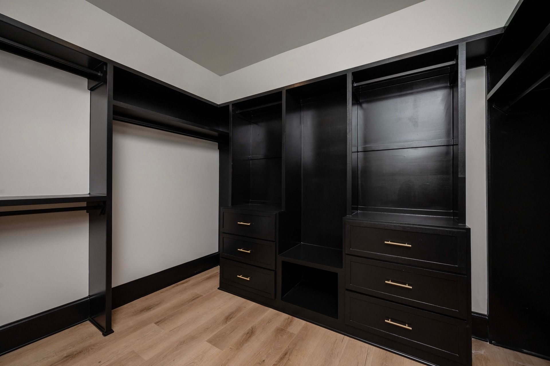 Black walk-in closet with drawers, shelves, and hanging rods. Light wood floor and white walls.