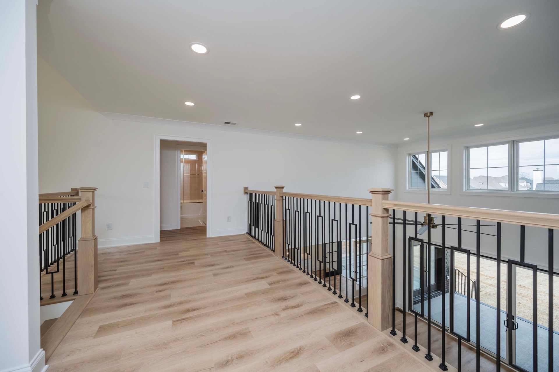 A bright, open upstairs hallway with a wooden floor, railing, and a doorway leading to a bathroom.
