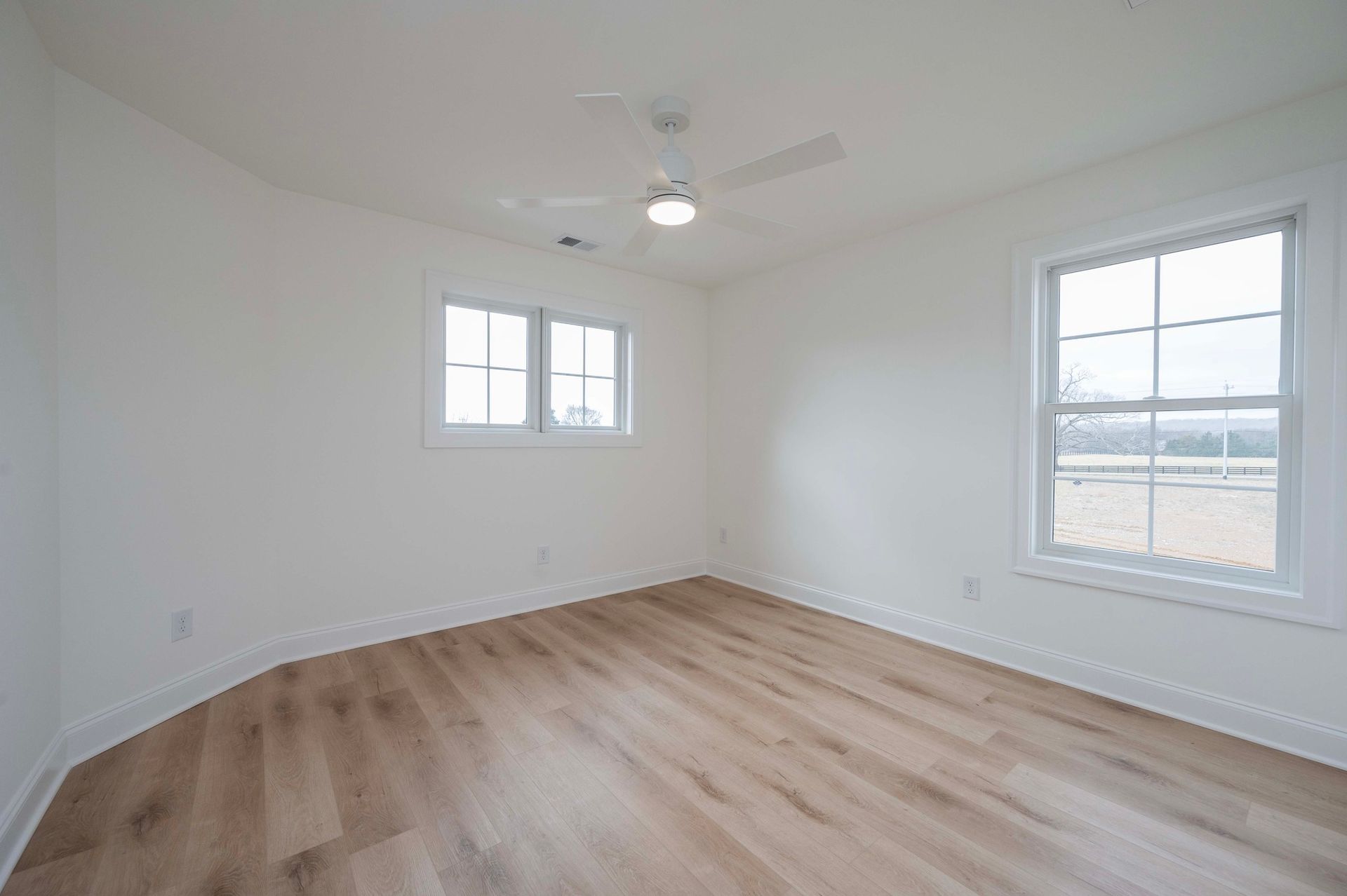 Empty bedroom with light wood floors, white walls, and two windows.