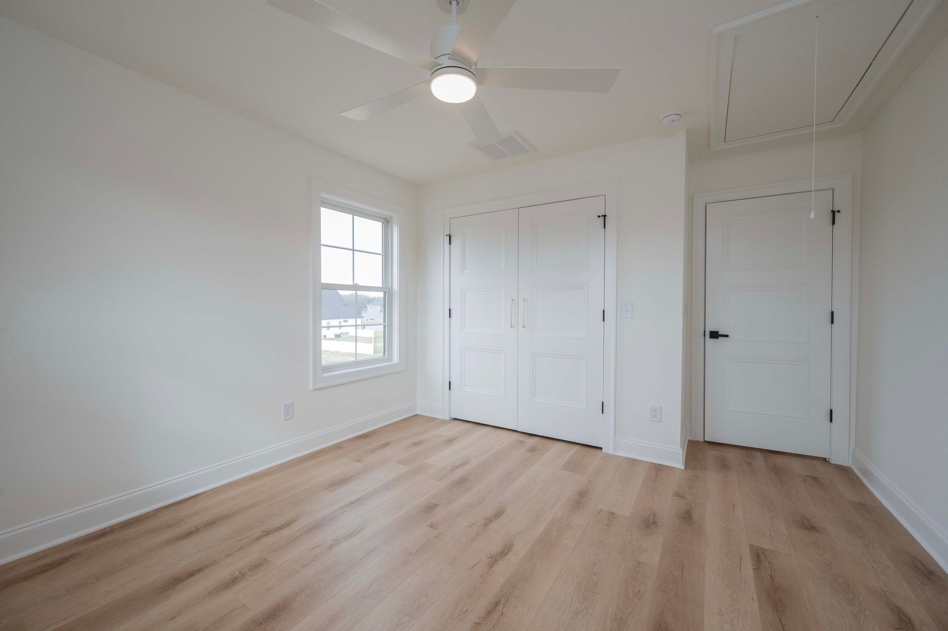 Empty bedroom with white walls, wood floor, window, closet, and door. Ceiling fan centered.