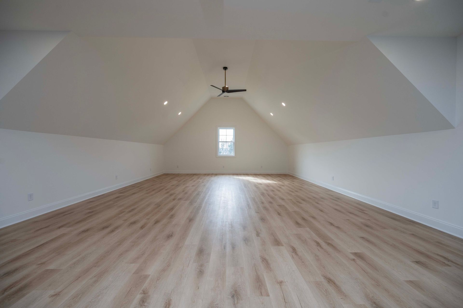 Empty, light-filled attic room with hardwood-style flooring, white walls, and a small window.
