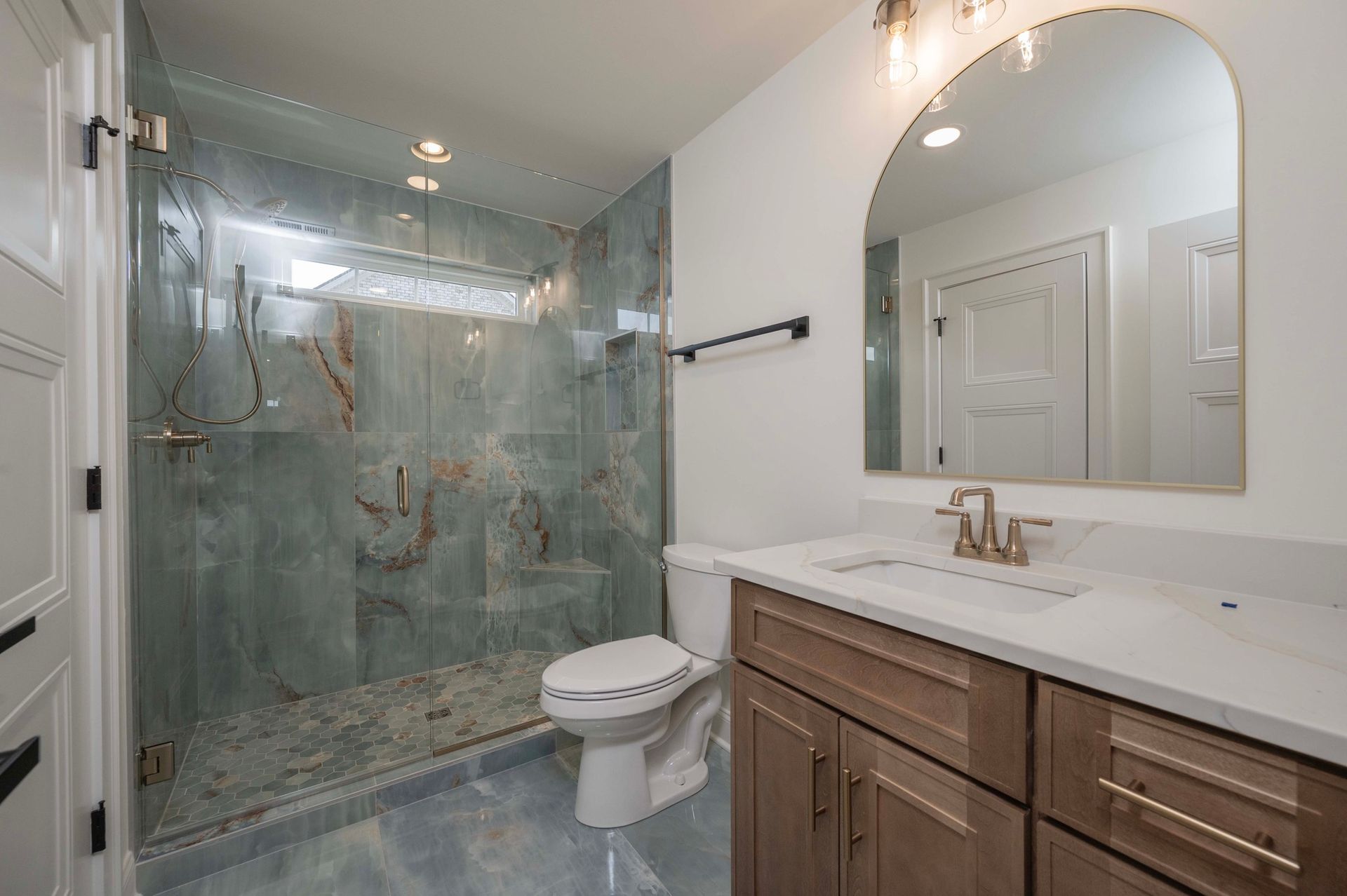 Bathroom with a glass shower, toilet, light-colored vanity, and large arched mirror.