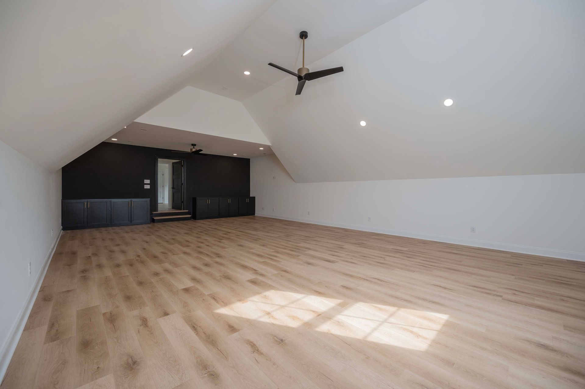 Empty room with angled white ceiling, wood floor, and black wall. Ceiling fan and recessed lighting.