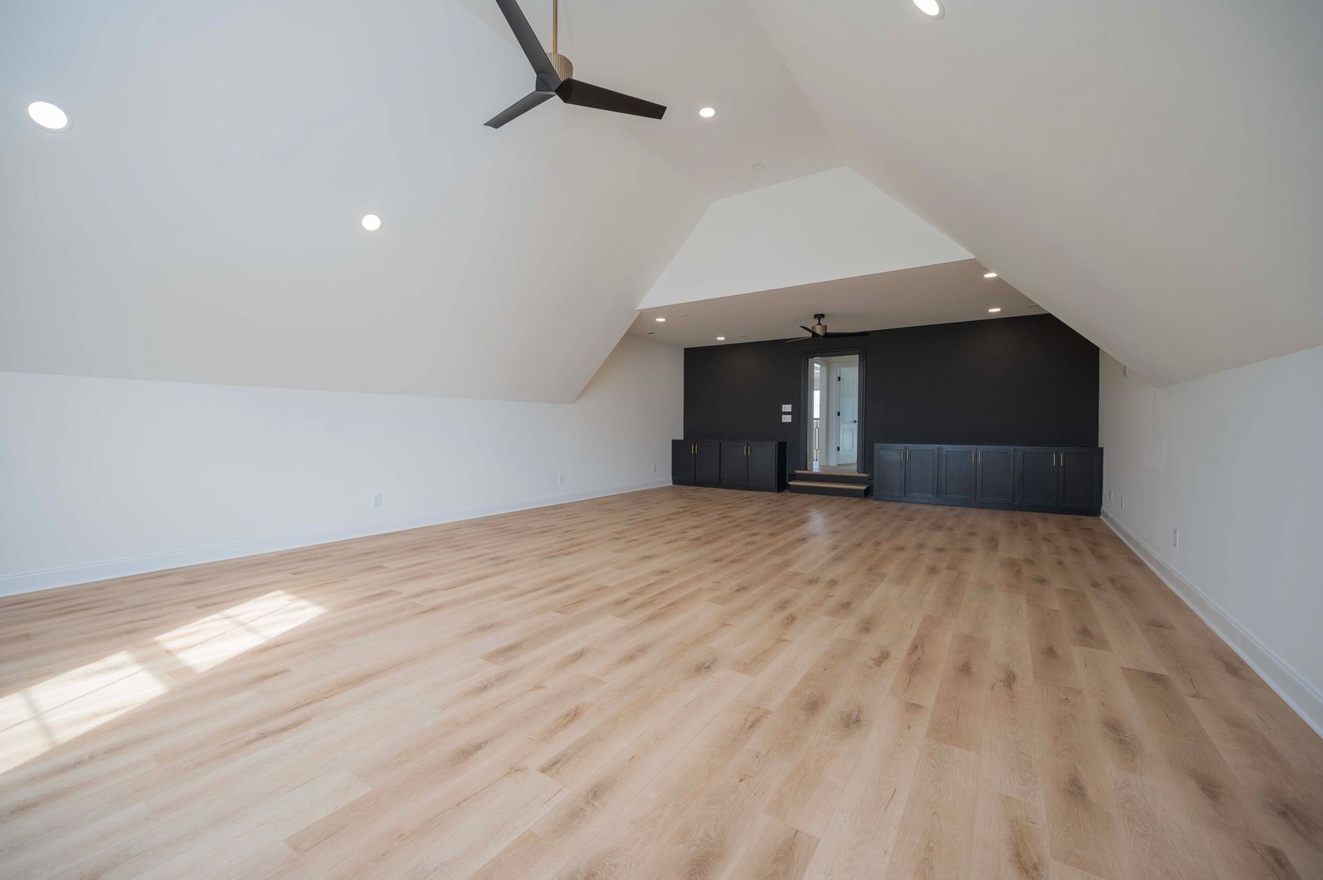 Spacious, empty room with light wood floors, white walls, and a black accent wall. Ceiling fan and recessed lighting.