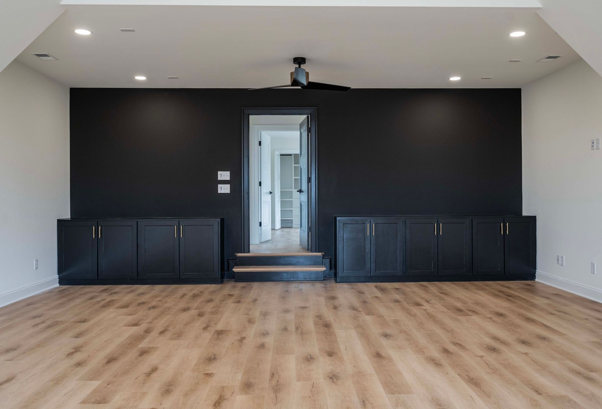 Large room with black accent wall, built-in black cabinets, and hardwood floors. Doorway leads to another room.