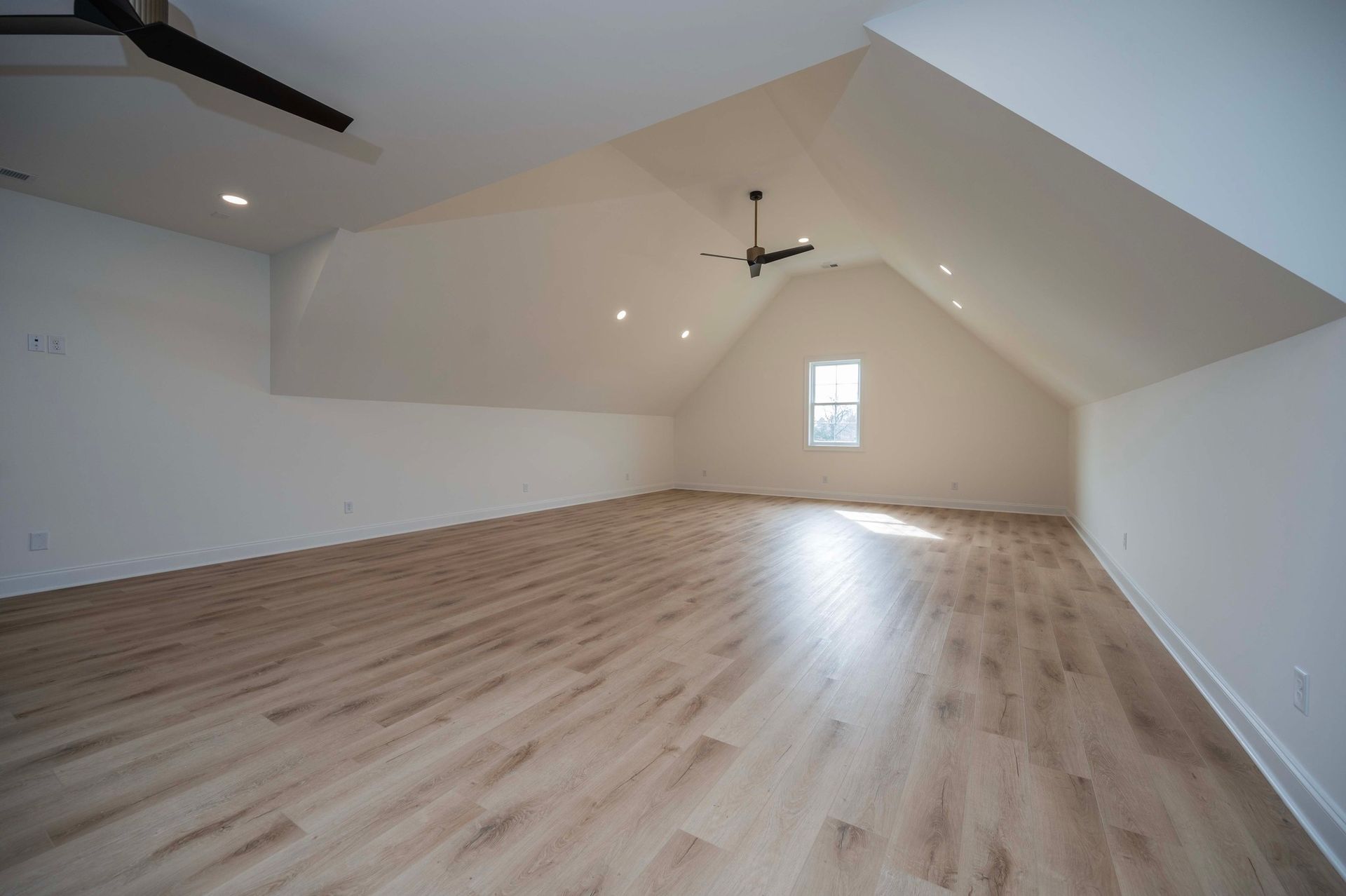 Empty attic room with wood flooring, white walls, and a small window.