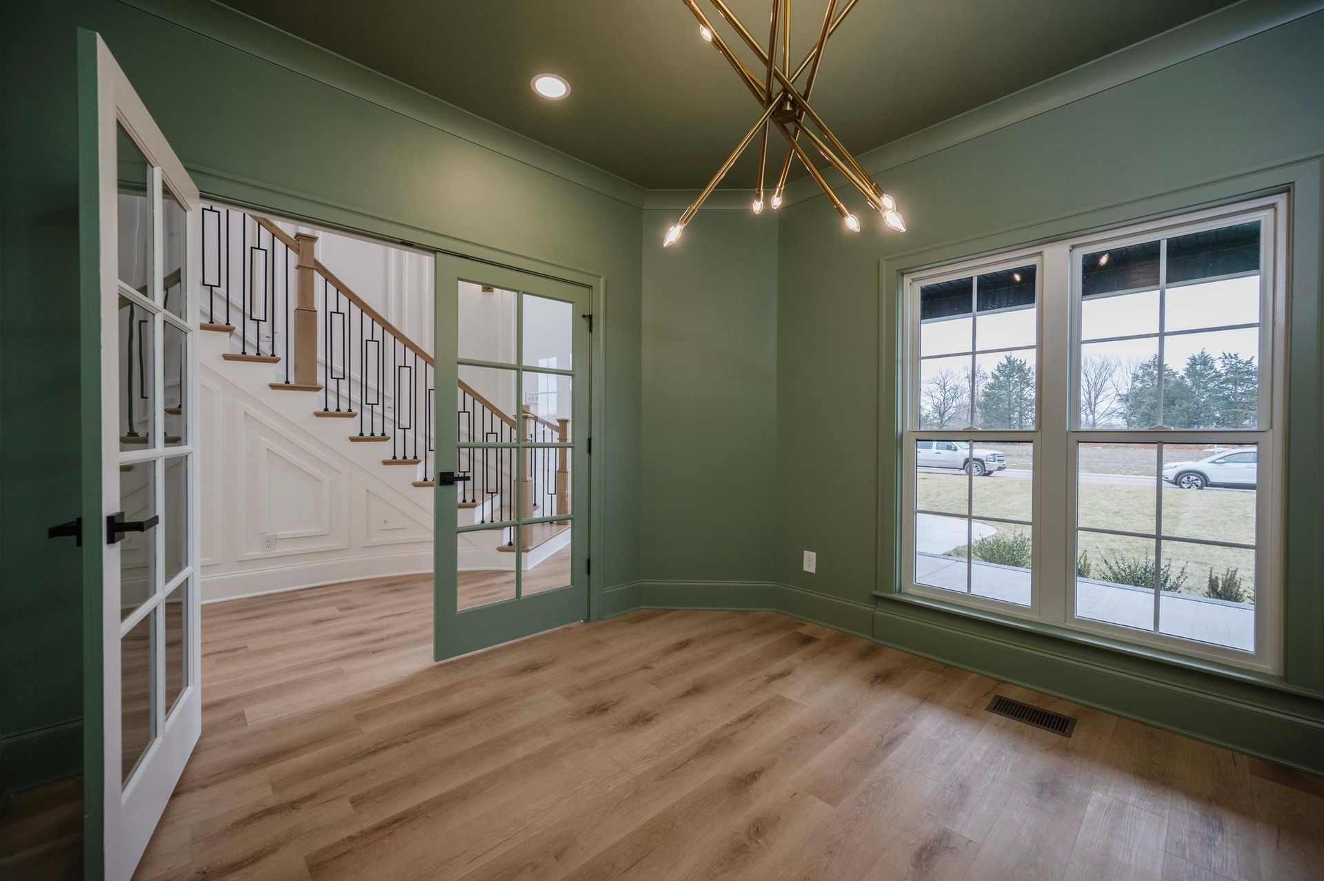 Green-walled room with glass-paneled doors, windows, and a hanging light fixture. Hardwood floors and a view of a staircase.