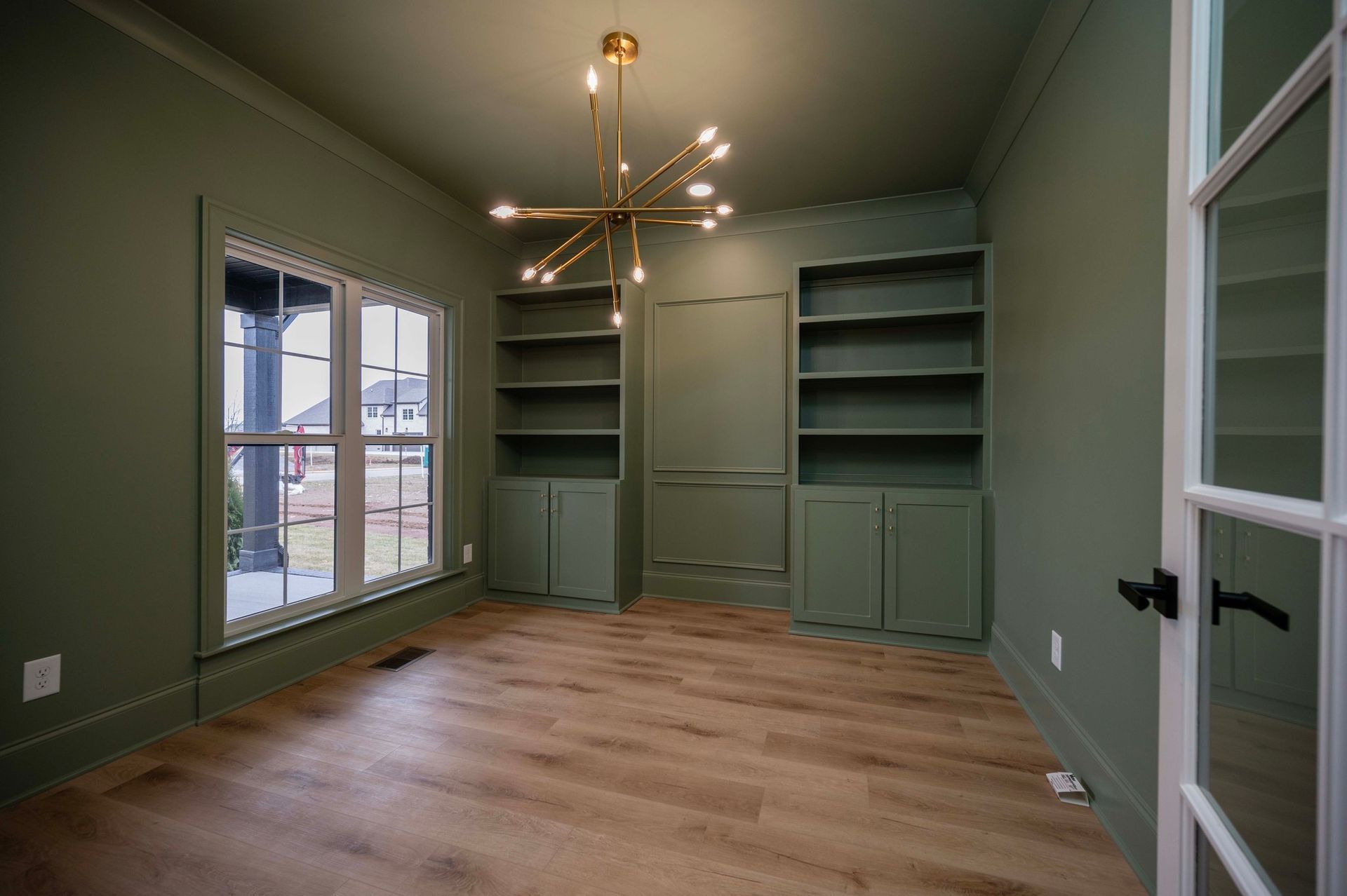 Green home office with built-in shelves, window, gold chandelier, and wood flooring.
