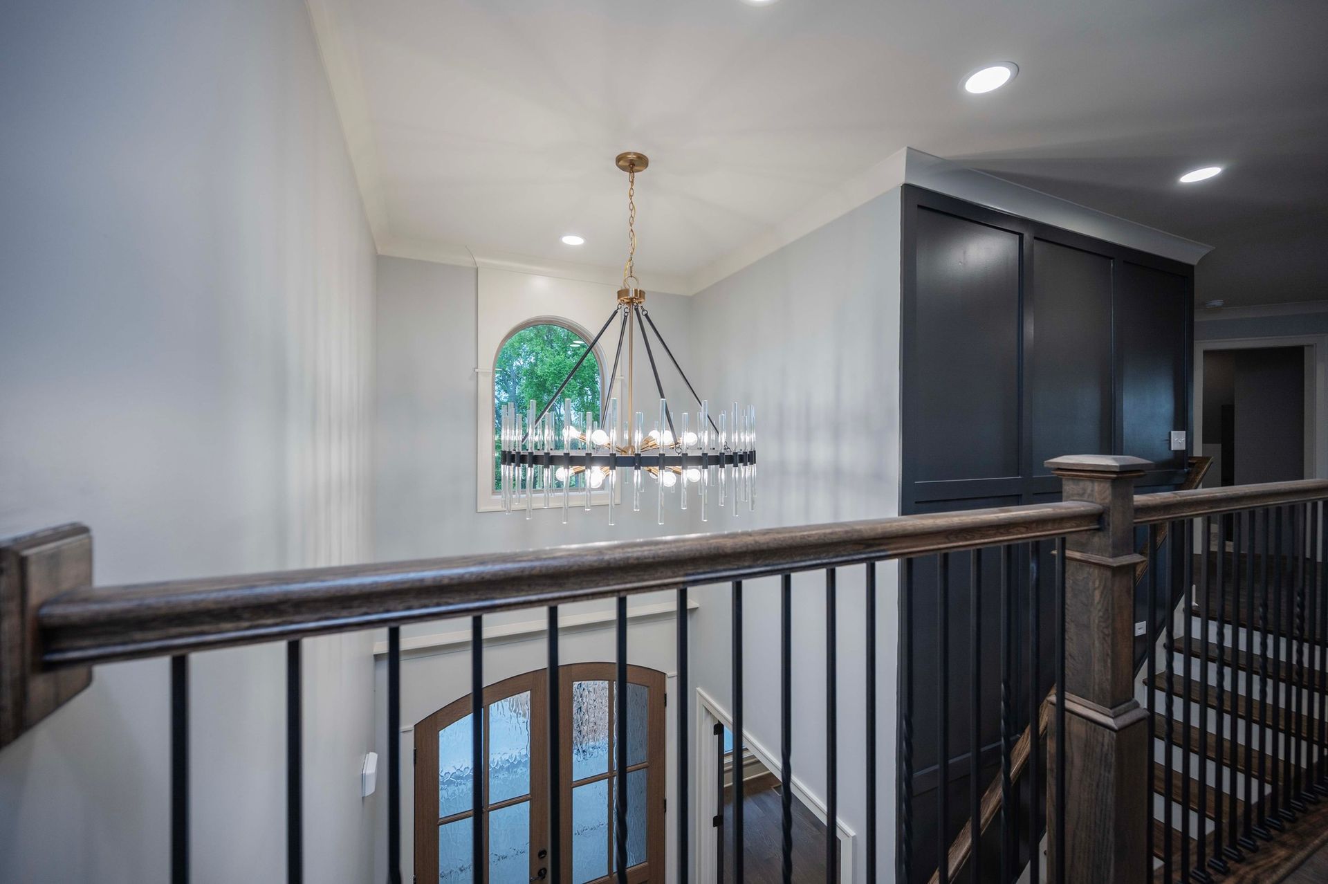 A hallway with a chandelier, window, and dark storage closet. Wooden railing and staircase are visible.