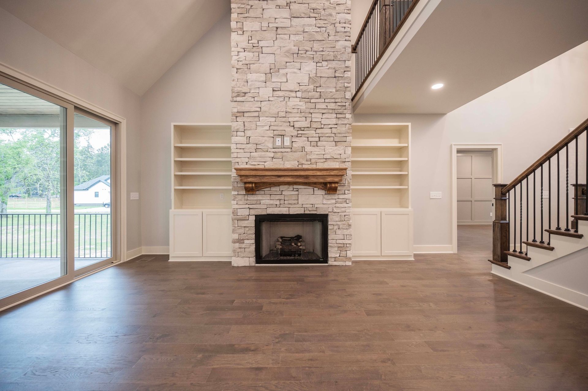 Living room with stone fireplace, built-in shelves, hardwood floor, and staircase.