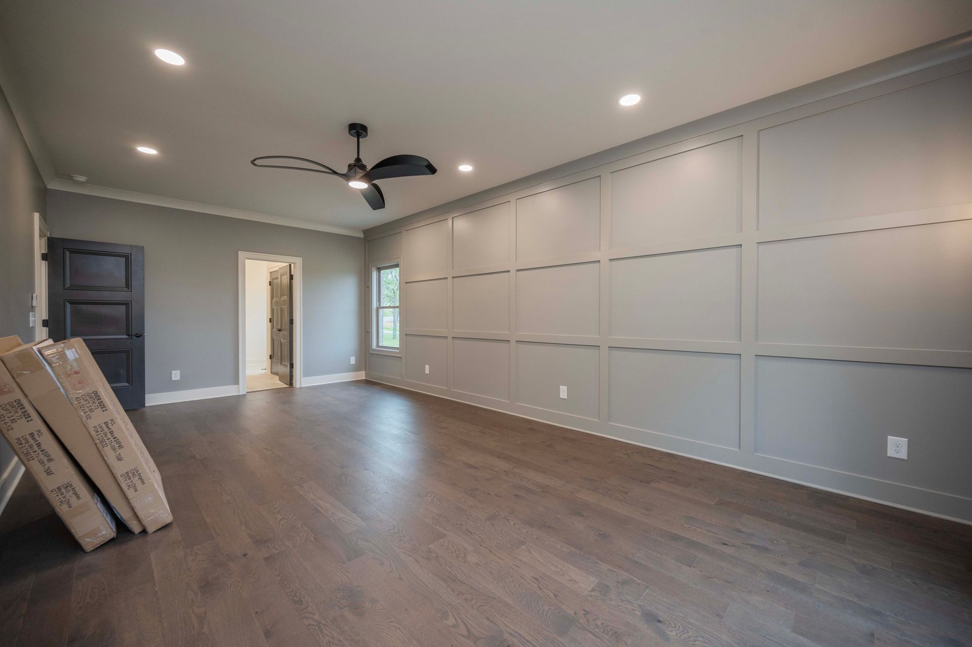 Empty, gray-walled room with dark wood floor, paneling, ceiling fan, and recessed lighting. Boxes lean against a dark door.