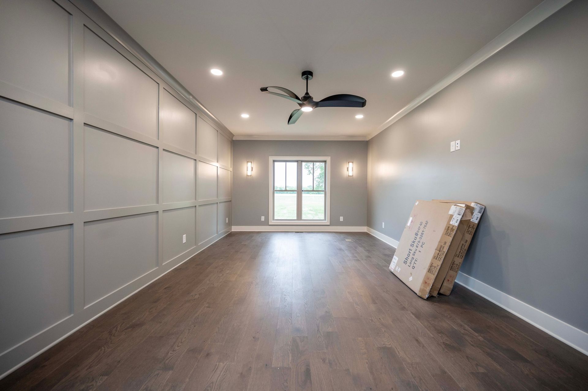 Empty room with gray walls, hardwood floor, and a ceiling fan. A box leans against the wall.