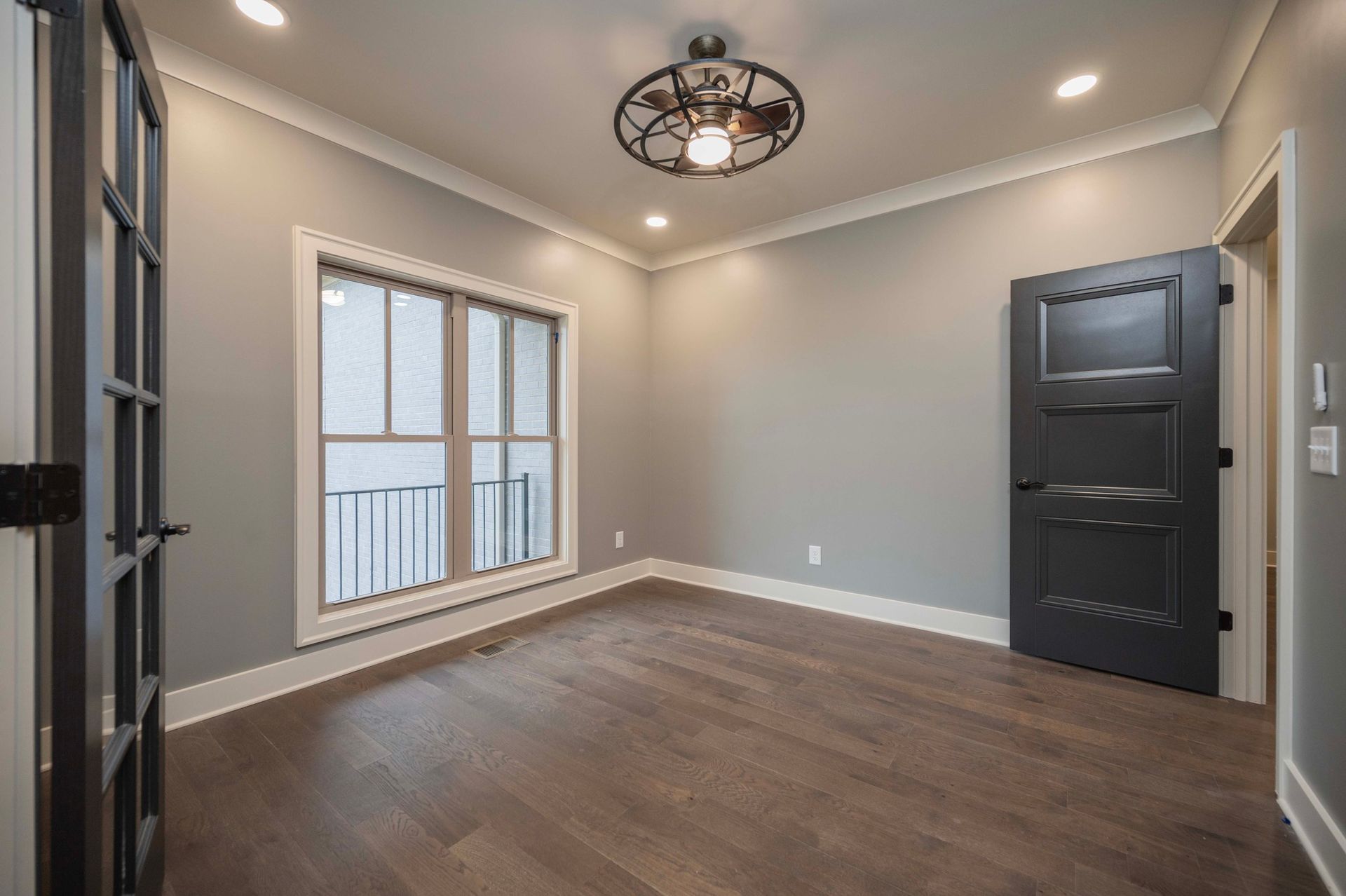 Empty room with gray walls, wood floors, a black door, and a window.