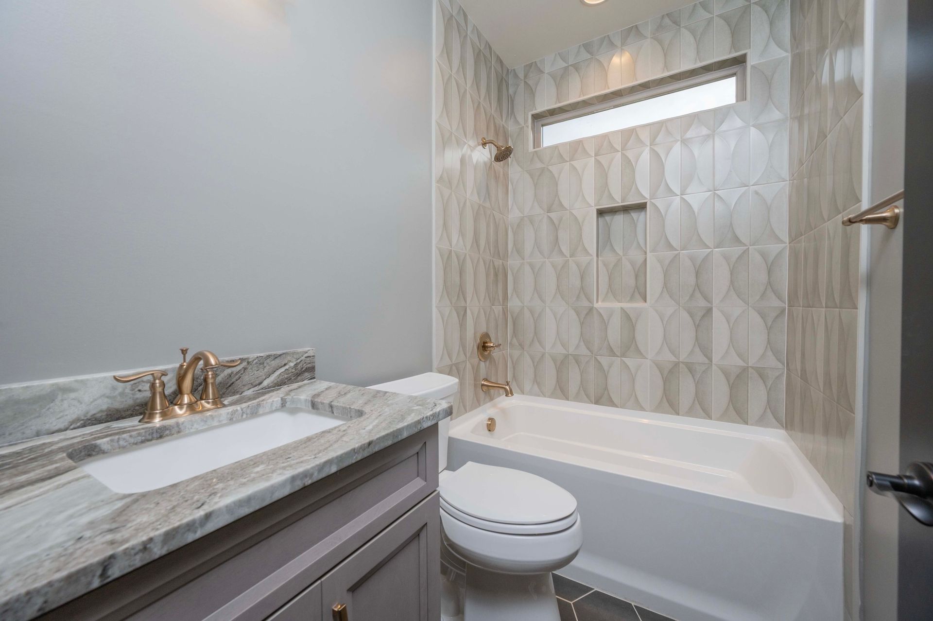 Bathroom with gray walls, patterned tile, white tub, toilet, and vanity with gold fixtures.