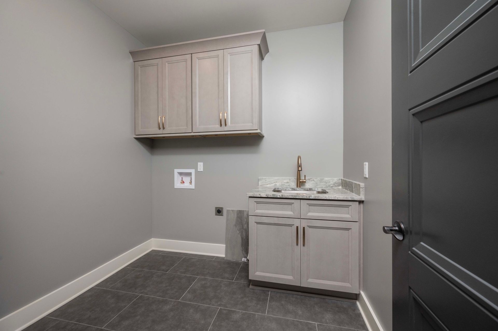 Laundry room with gray walls, cabinets, and a sink. Dark floor and door.