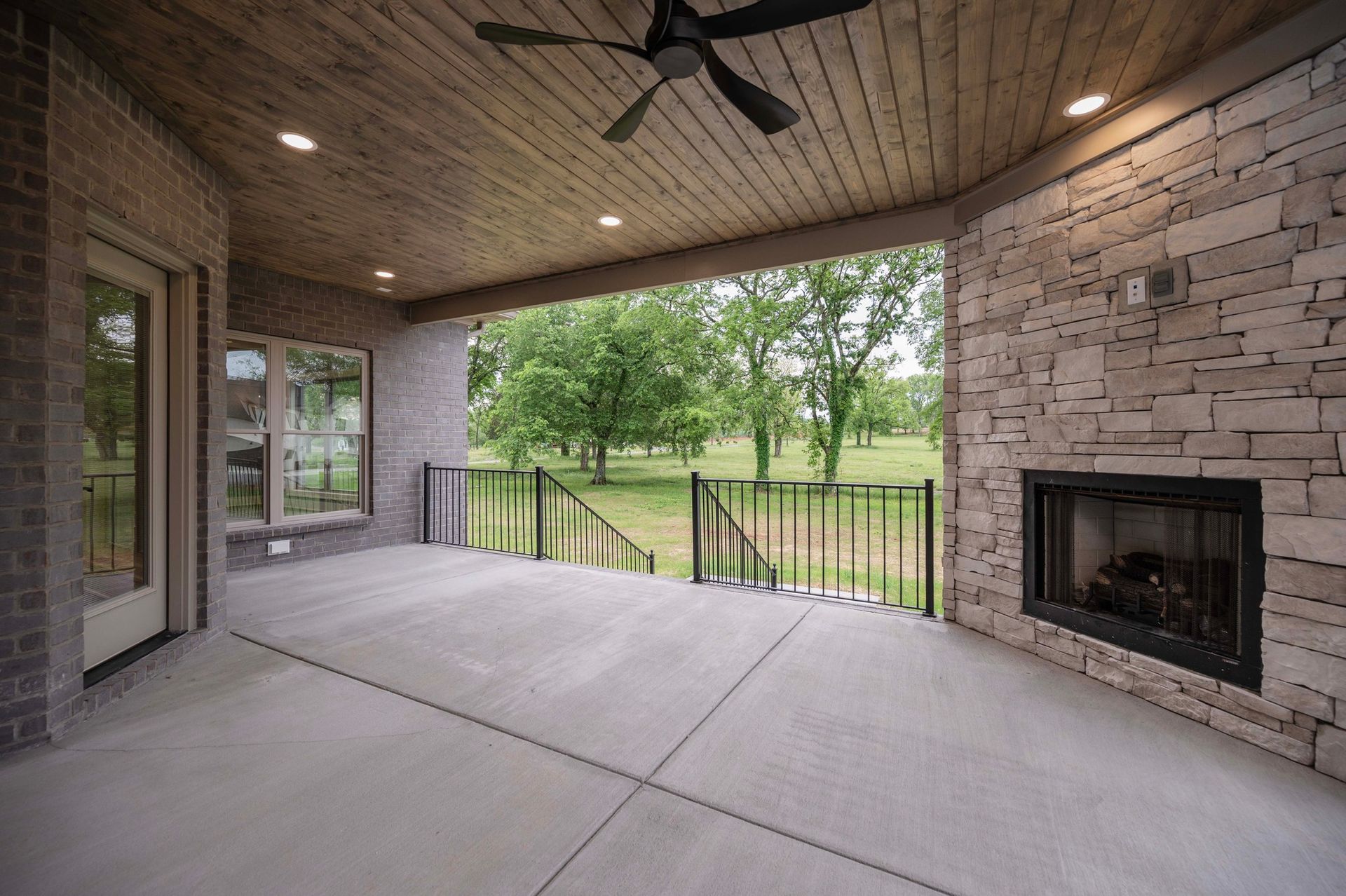 Covered patio with fireplace, stone wall, concrete floor, and view of green trees and yard.