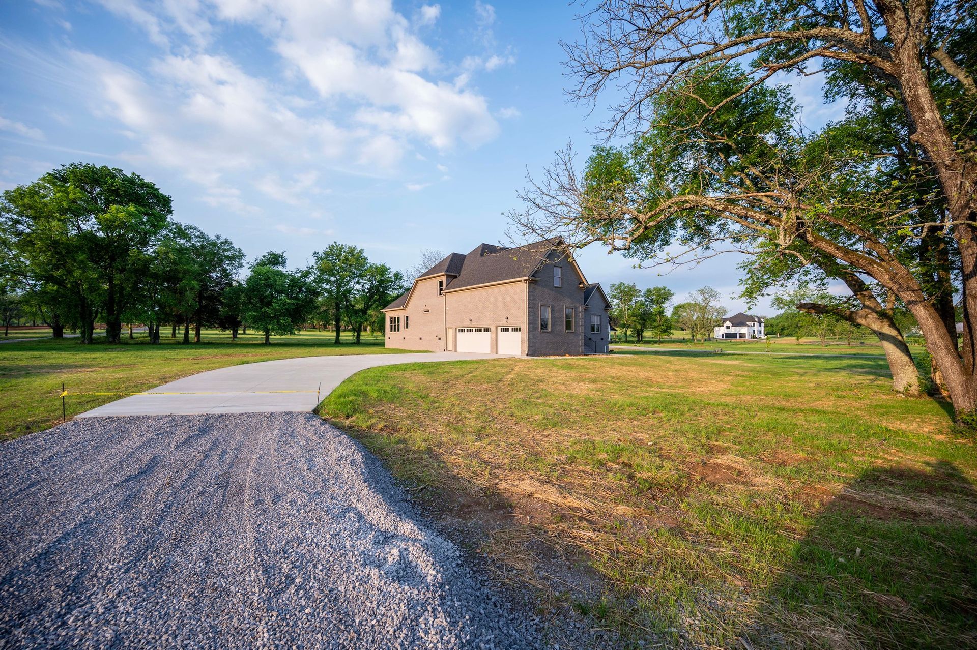 A gravel driveway leads to a two-story beige house with a brick facade in a grassy field, under a sunny sky.