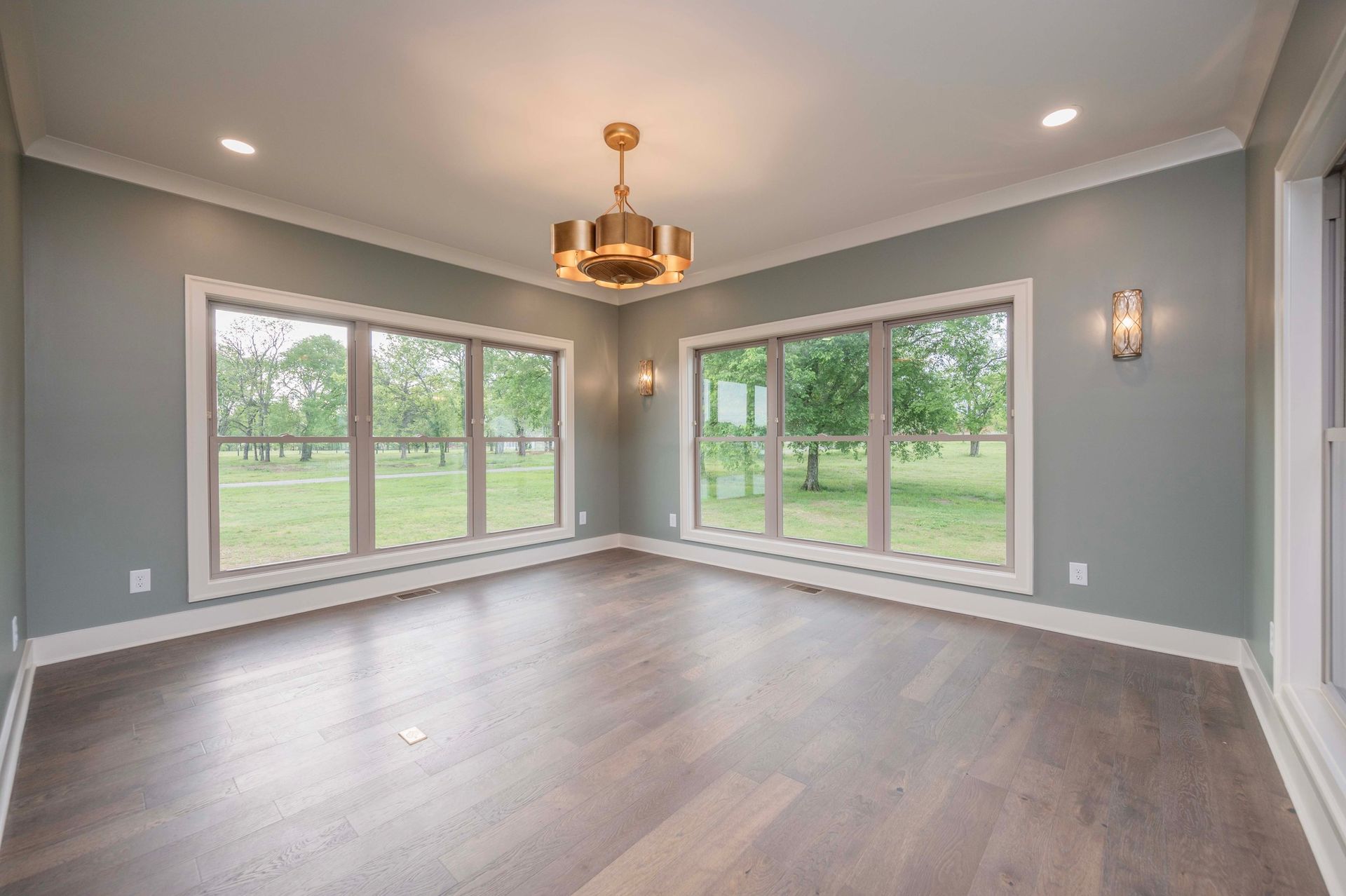 Empty room with gray walls, large windows, wood floor, and a gold chandelier.