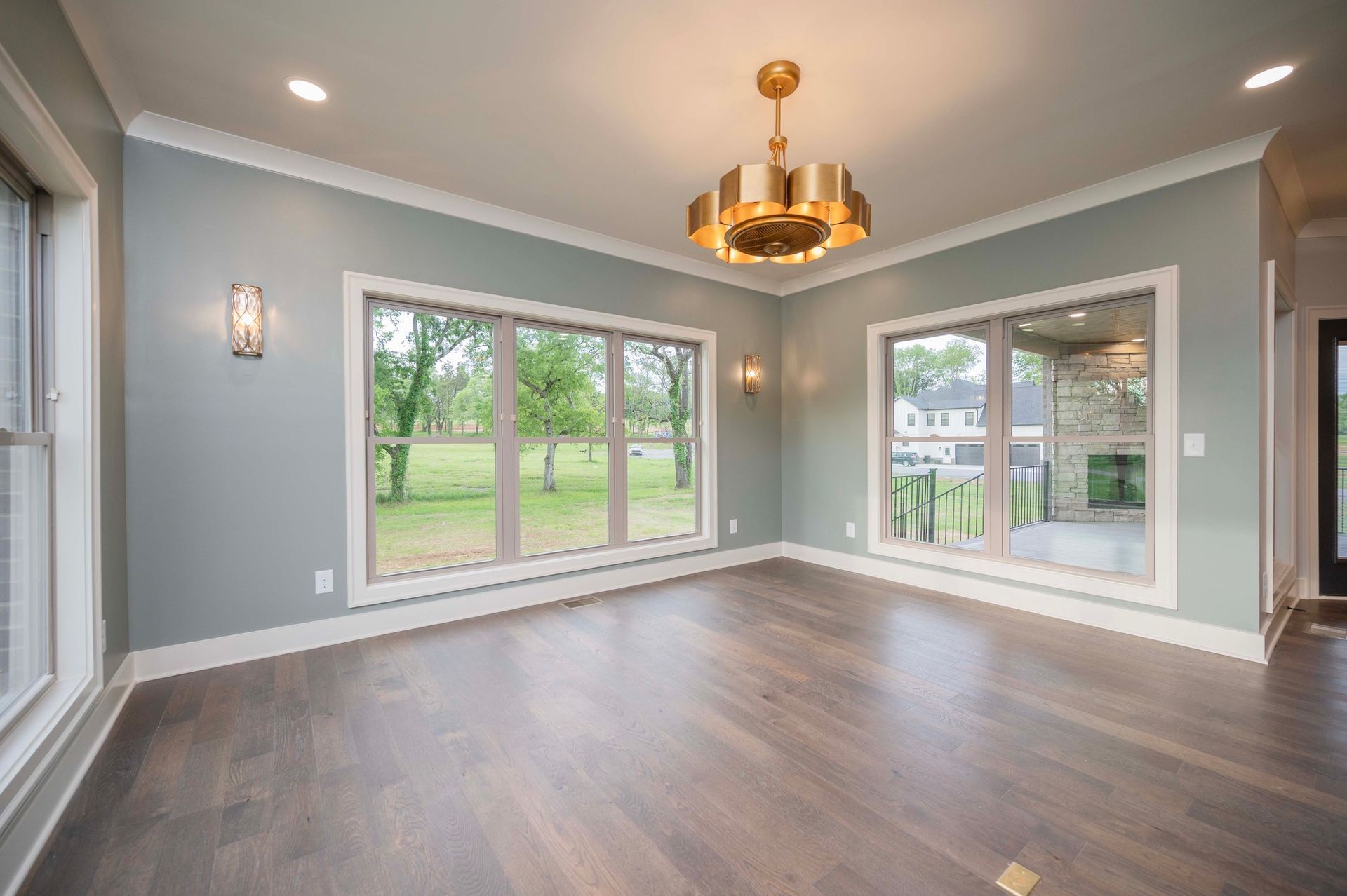 Empty room with light blue walls, wooden floors, large windows, and a gold chandelier.