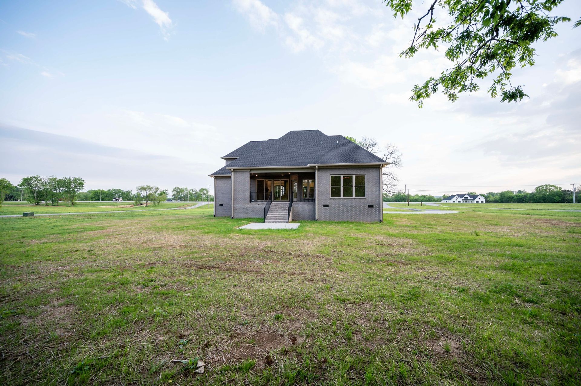 Gray house with dark roof on a large grassy lot, trees in background, cloudy sky.