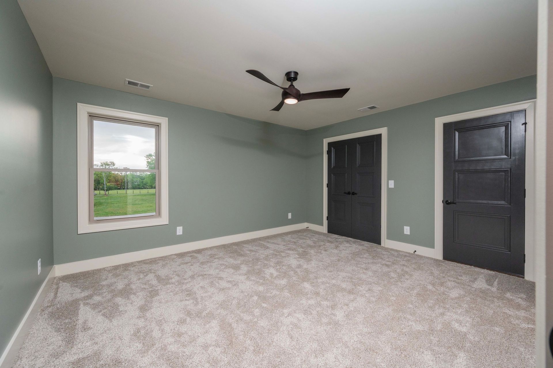 Empty bedroom with gray carpet, teal walls, black doors, and a window overlooking greenery.