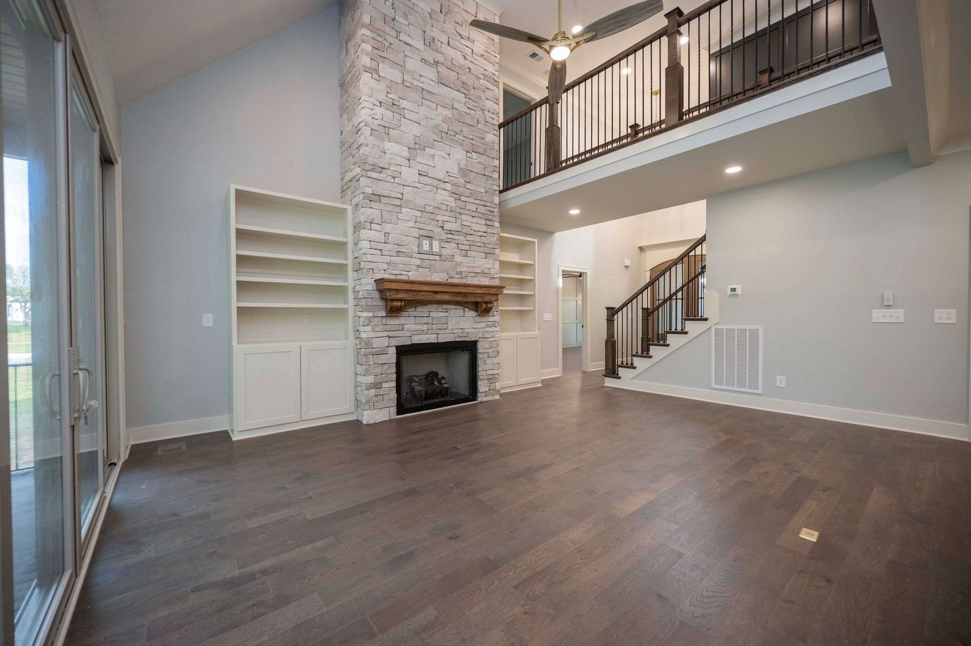 Spacious living room with gray walls, wood floors, and a brick fireplace. Features a second-floor balcony and built-in shelves.