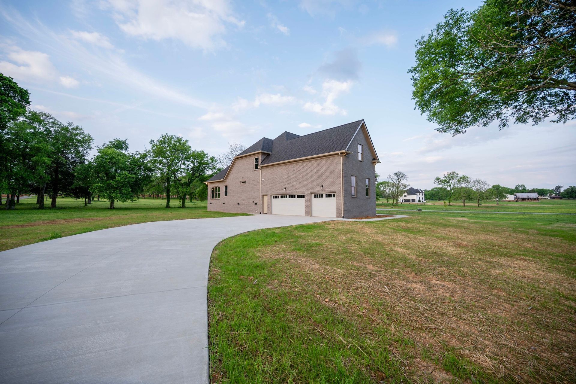 A light brick house with a two-car garage, long concrete driveway, and grassy lawn under a cloudy sky.