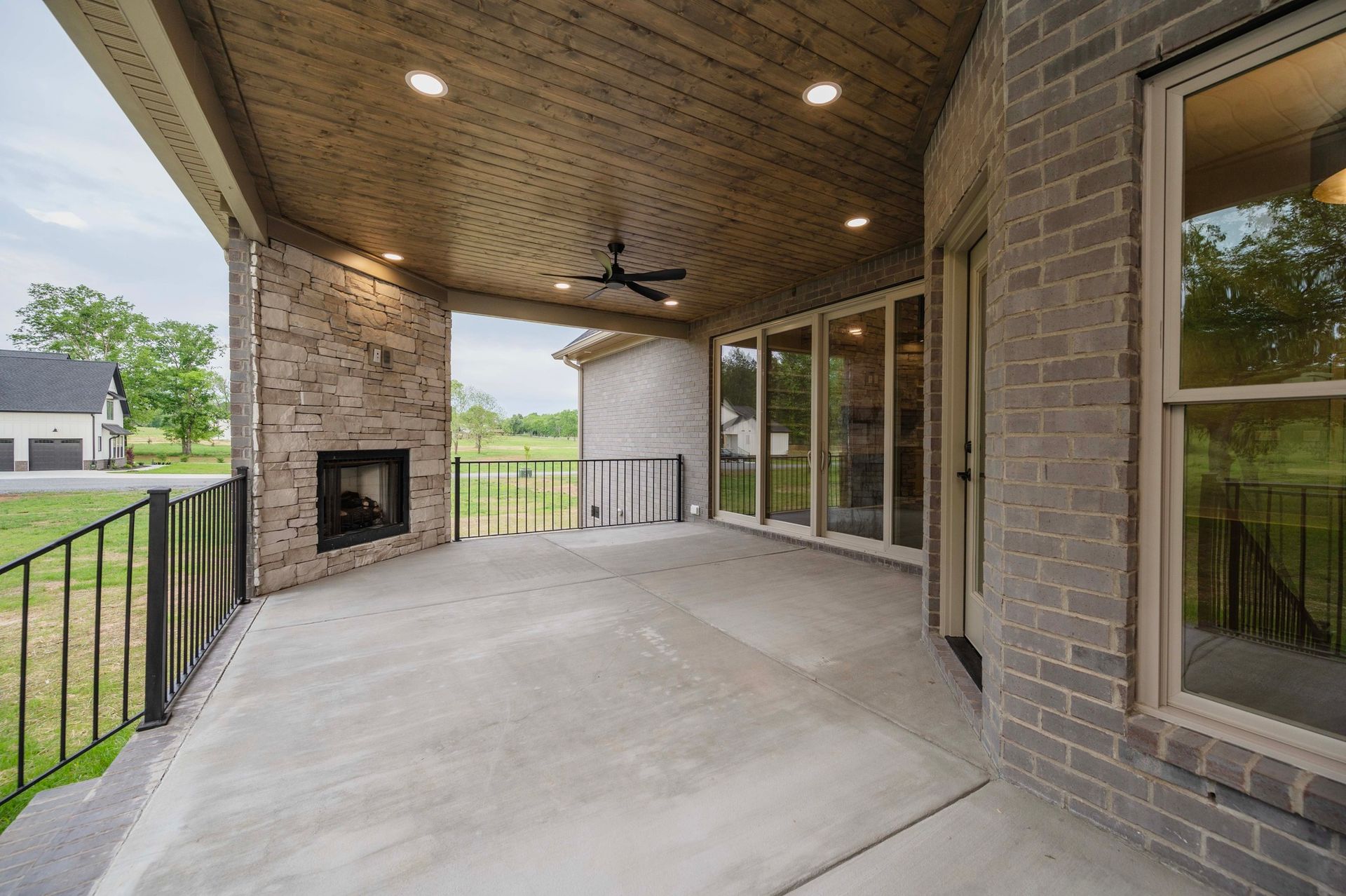 Covered outdoor patio with fireplace, brick wall, and glass doors; black railing and ceiling fan.