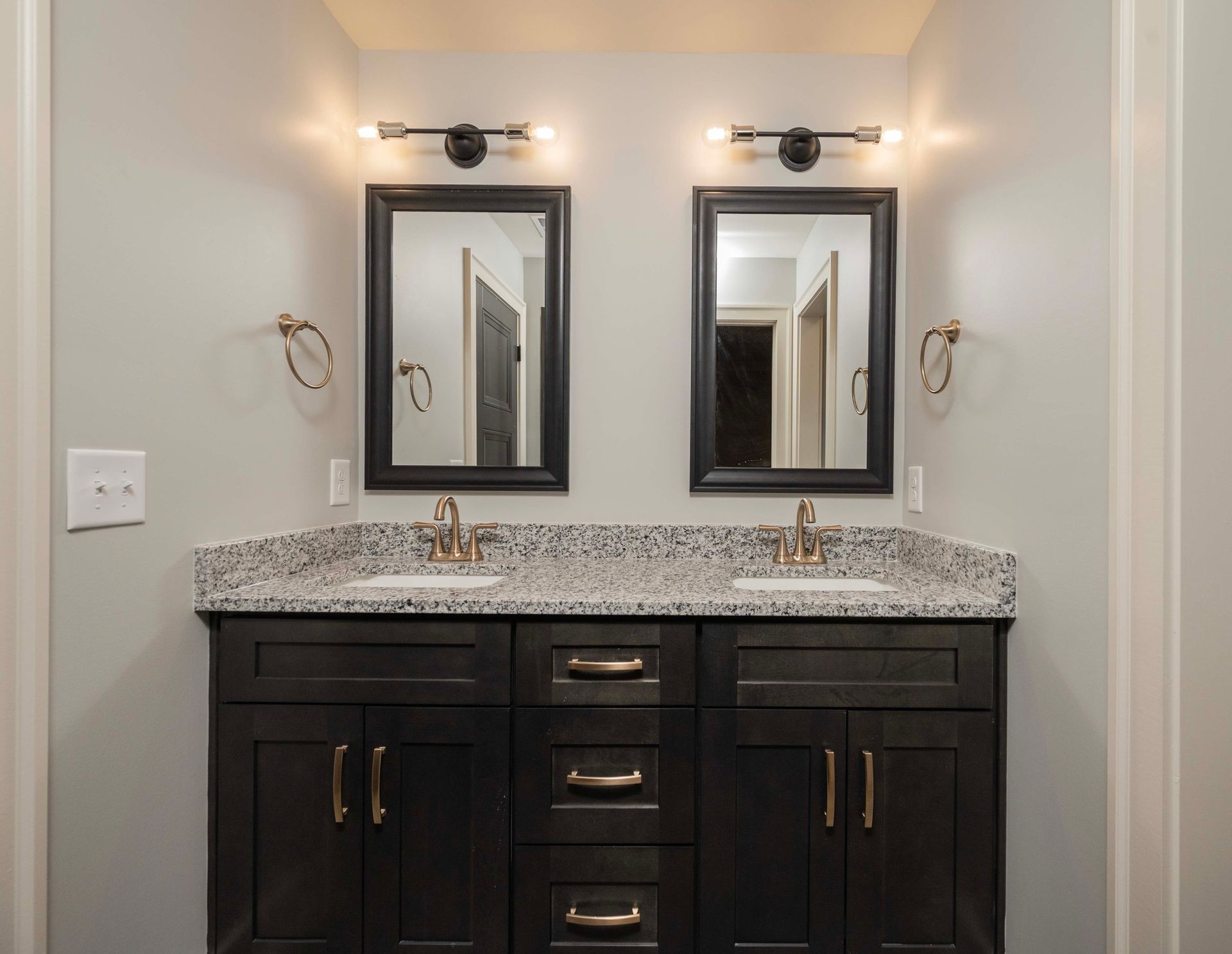 Dual vanity bathroom with dark cabinets, speckled countertop, two mirrors, and gold fixtures.