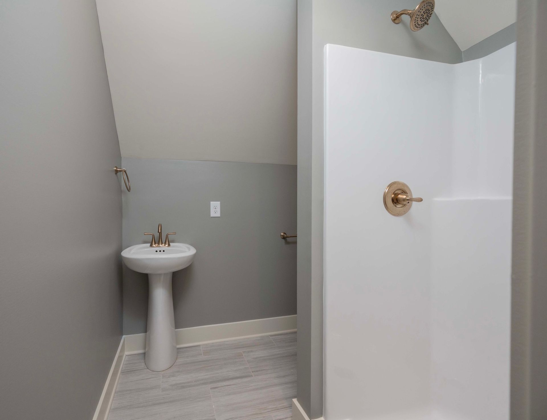 Bathroom with pedestal sink, shower, and angled ceiling. Gray walls and flooring, white fixtures.