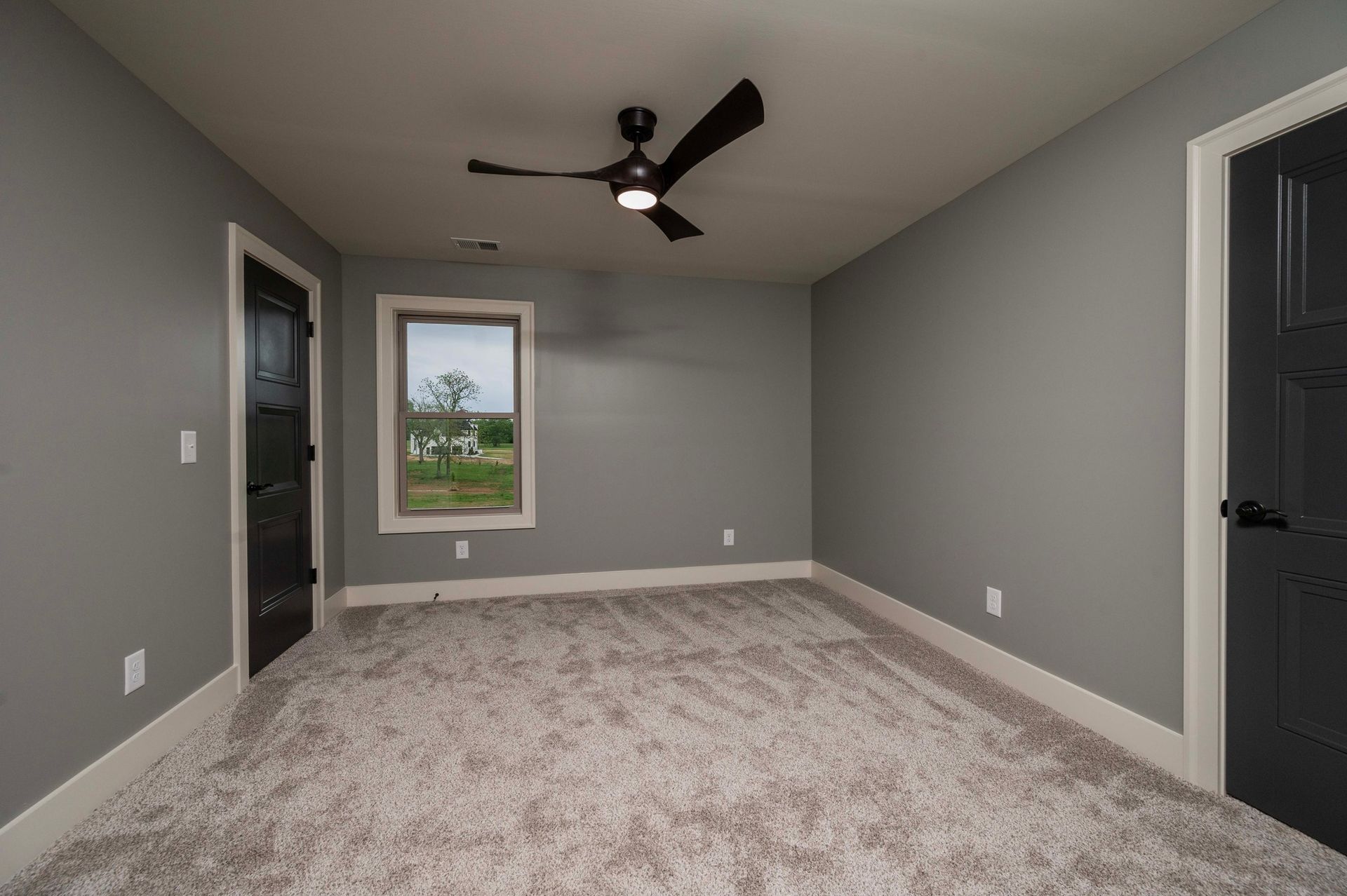 Empty room with gray walls, black doors, a window, and a dark ceiling fan; beige carpet.