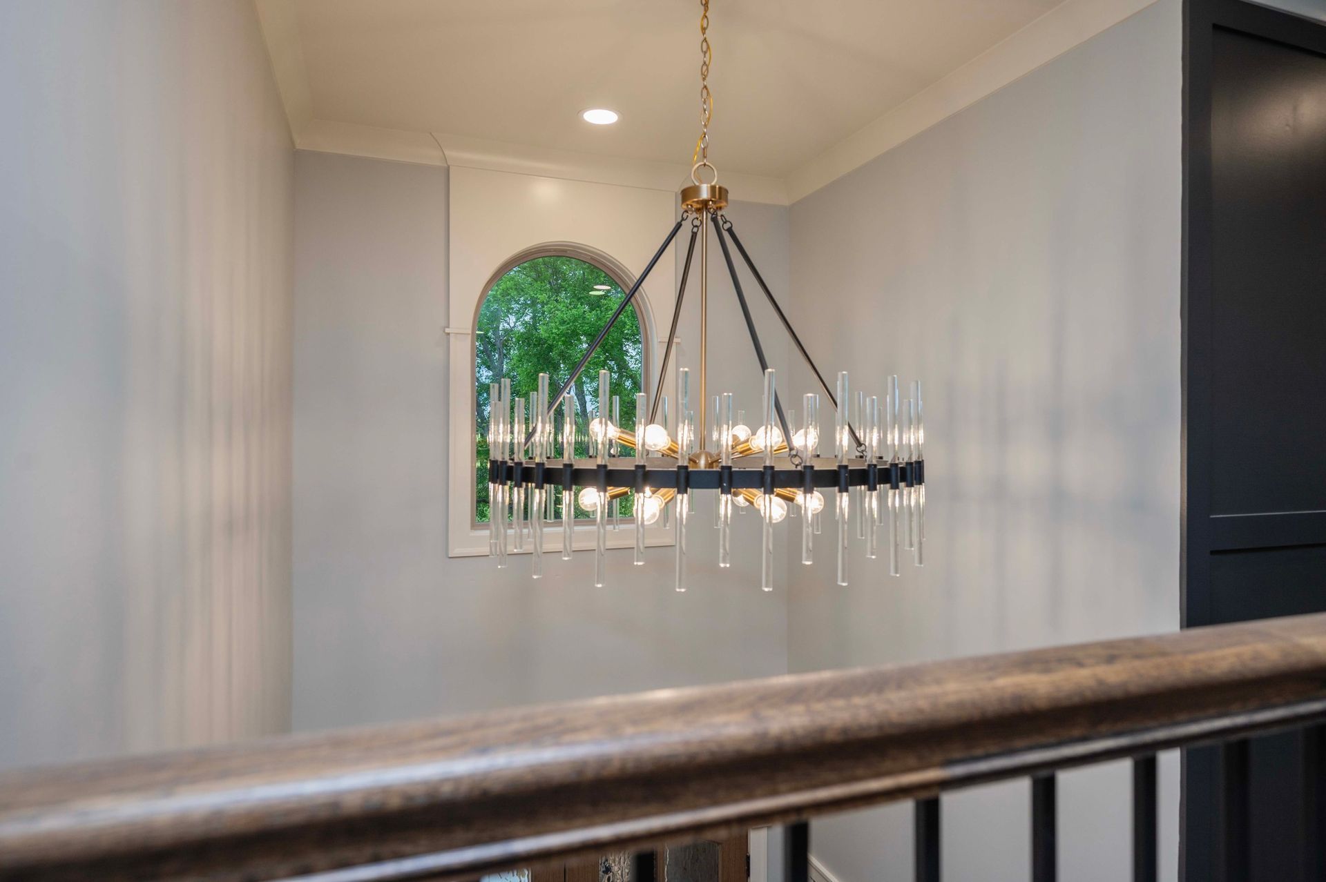 Hallway with a chandelier and arched window, overlooking greenery.