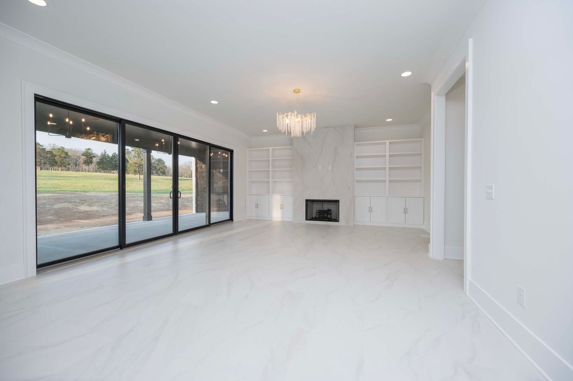 Empty white living room with large glass doors overlooking a green field. Fireplace and built-in shelves are present.