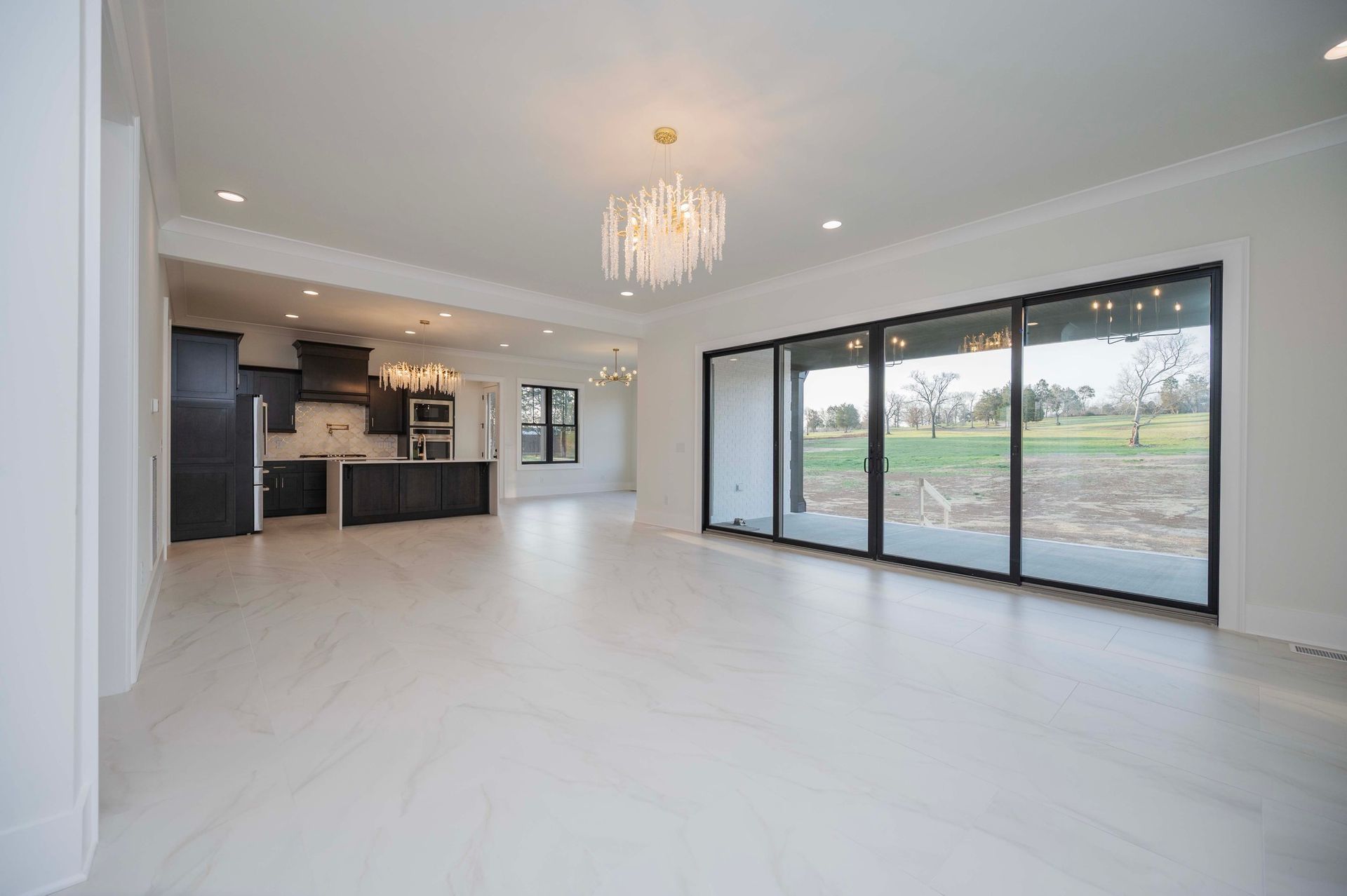 Bright, empty room with large sliding glass doors overlooking a green field. A dark kitchen is visible.