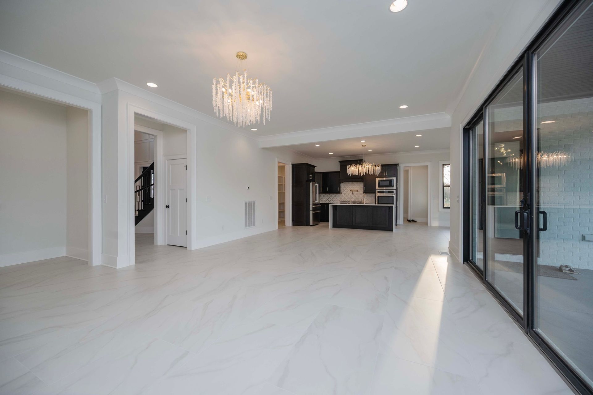 Bright, empty open-concept living area with white tile floors, chandelier, and black framed sliding glass doors to exterior.