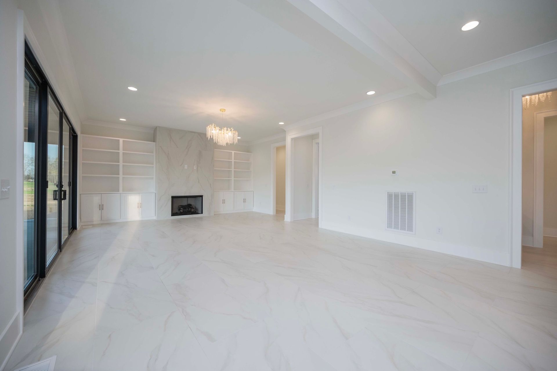 Empty, bright, white-walled living room with large sliding glass door. Fireplace, built-in shelving, and a chandelier are visible.