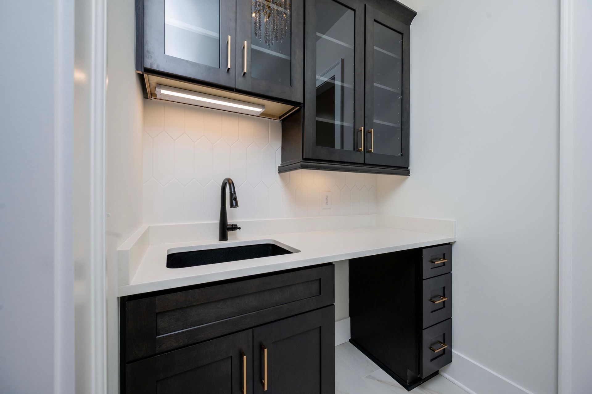 Black cabinetry with sink and countertop in a butler's pantry, white walls, and glass-front upper cabinets.
