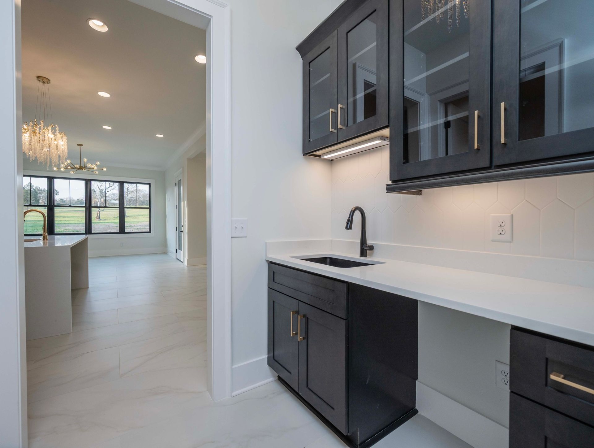 Dark-cabinet bar area with a sink, white countertop, and glass-front upper cabinets, leading into a bright white room.