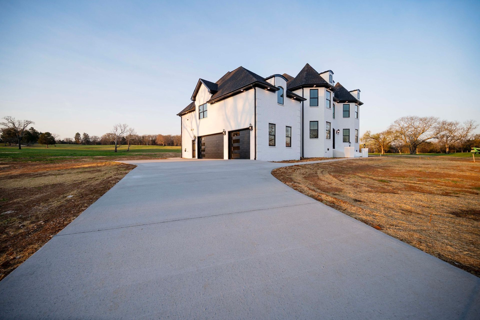 White two-story house with black trim, driveway, and garage doors. Set in a grassy field under a blue sky.