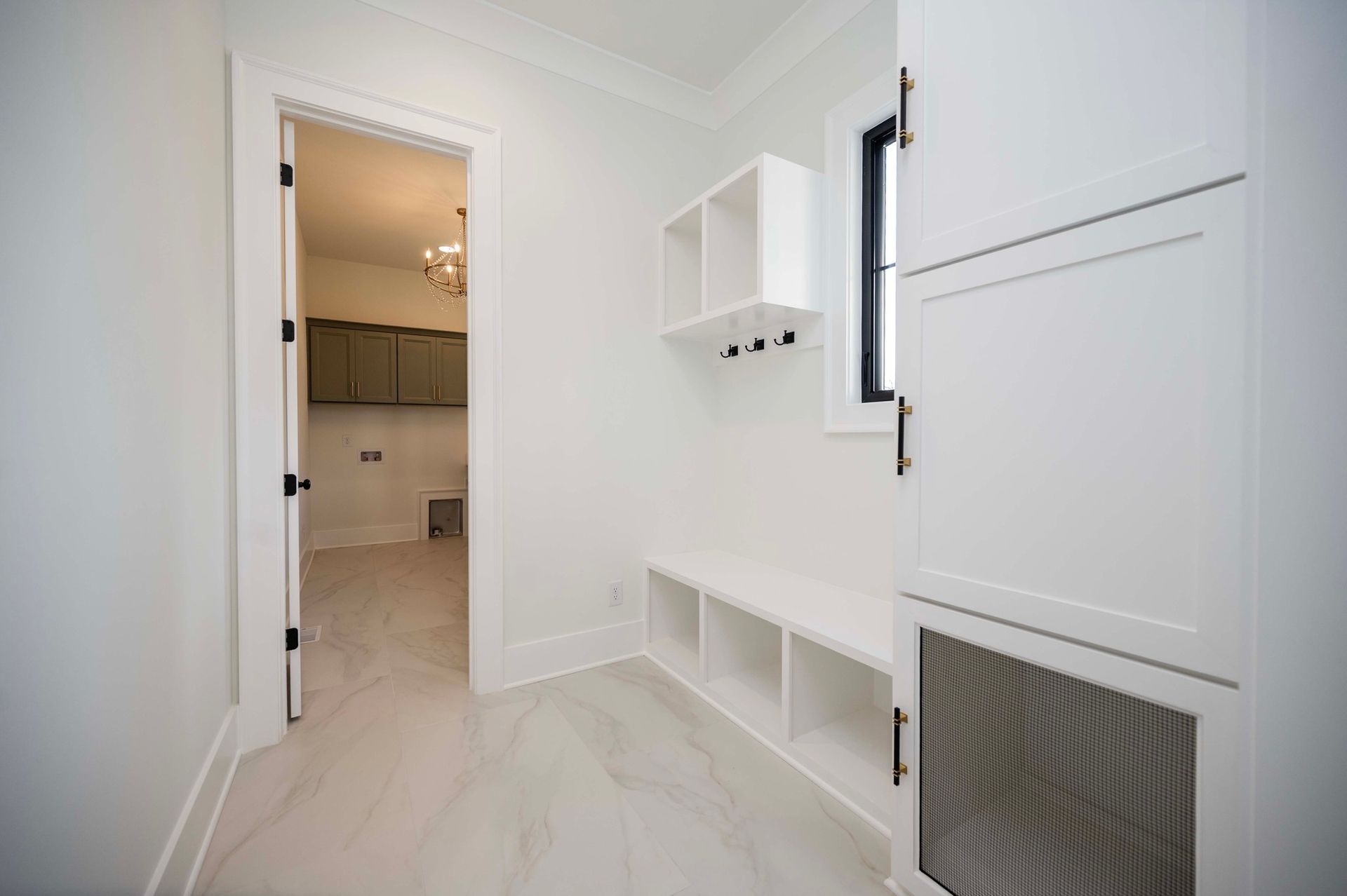 White-walled laundry room with built-in storage, bench, open doorway to more room, and a small window.