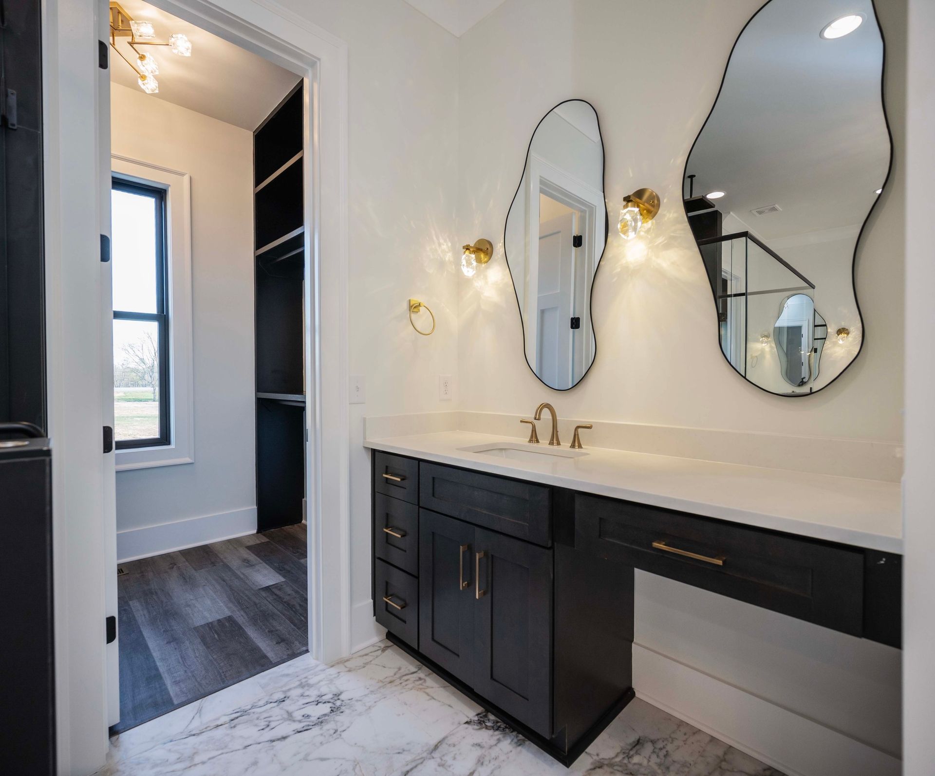 Vanity area with black cabinets, wavy mirrors, and gold sconces. Closet in the background.