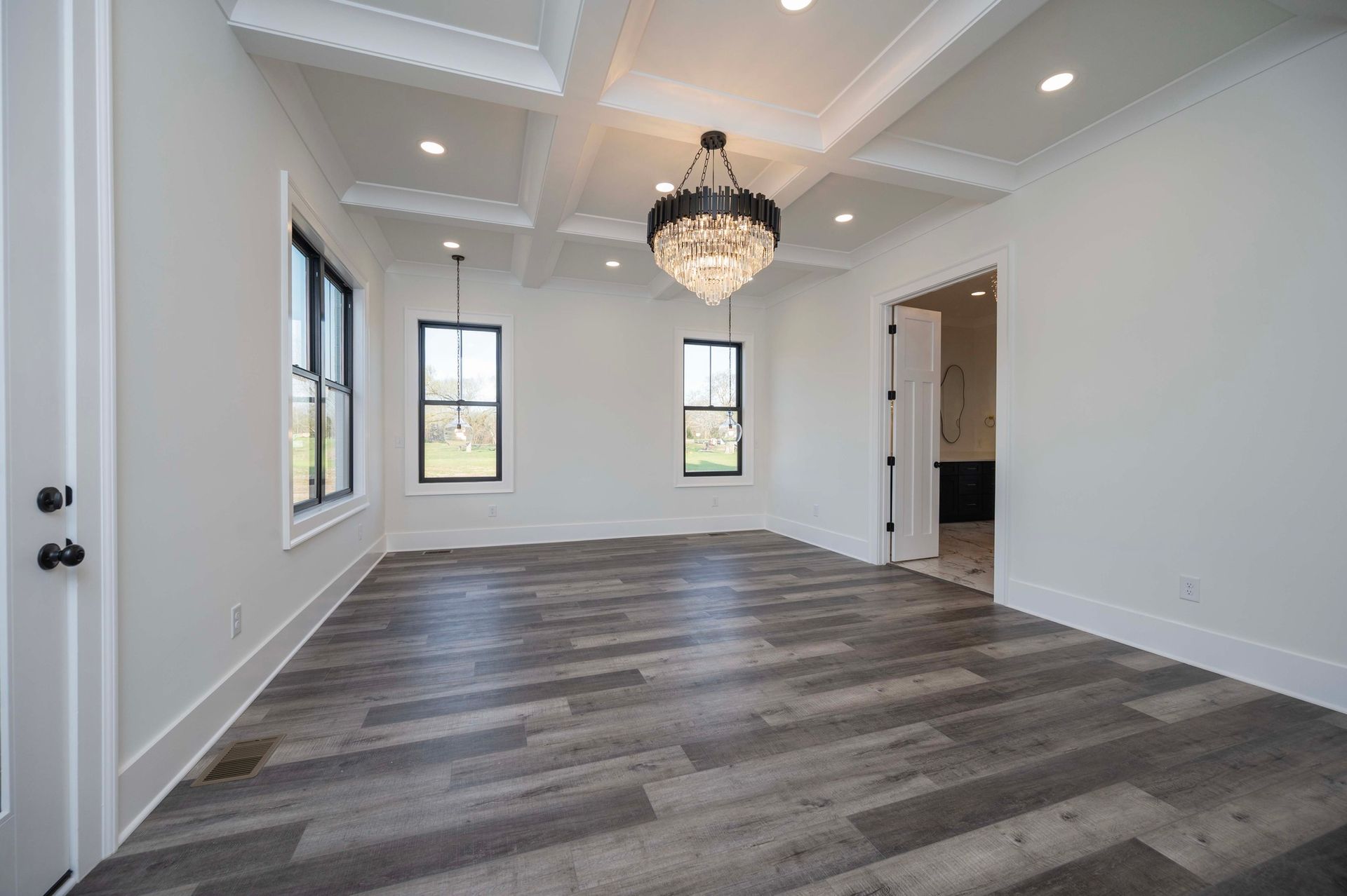 Empty room with dark wood-look flooring, white walls, and a chandelier; three windows and a door are visible.