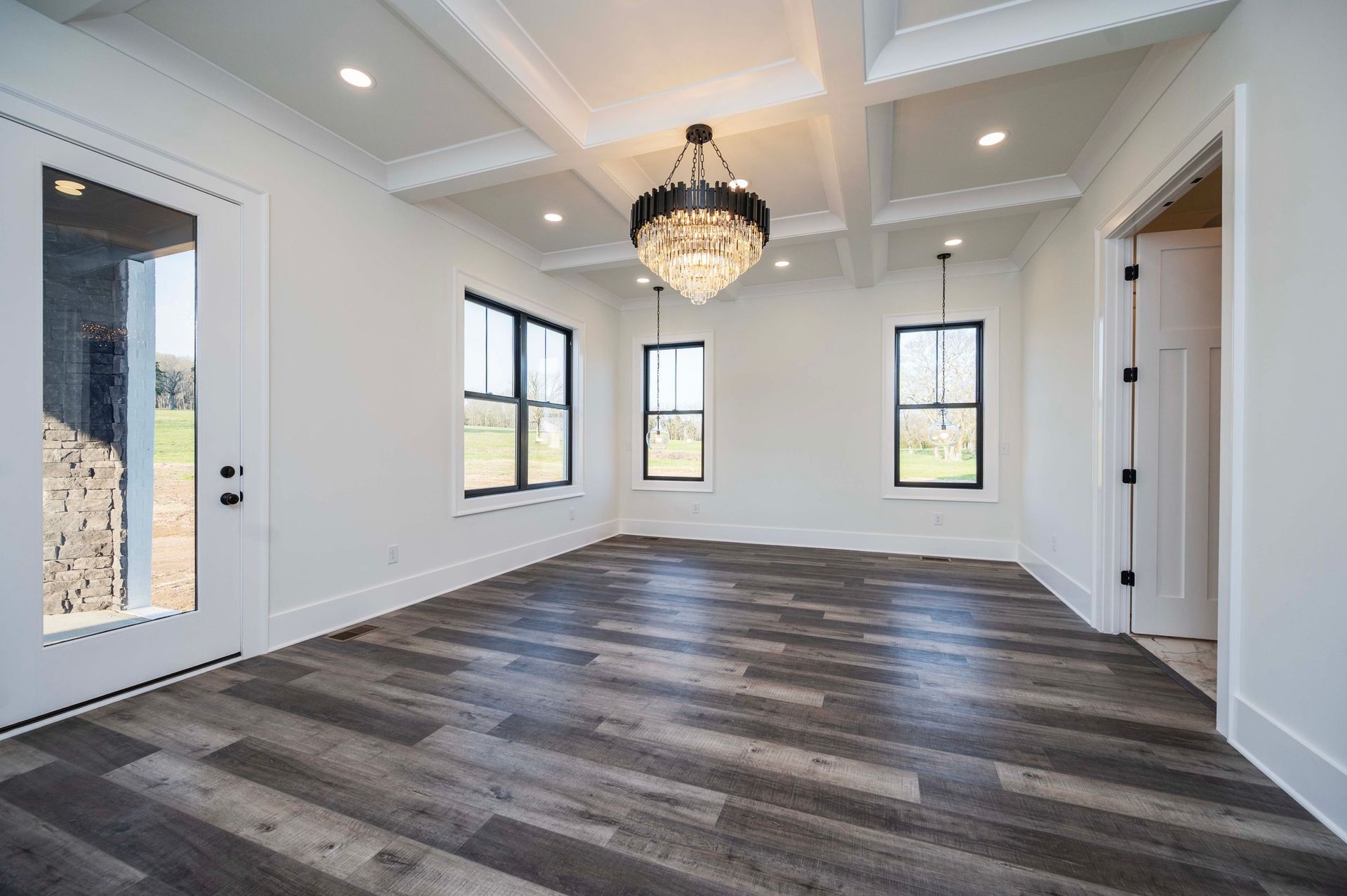 Empty room with hardwood floors, white walls, black-framed windows, and a chandelier.