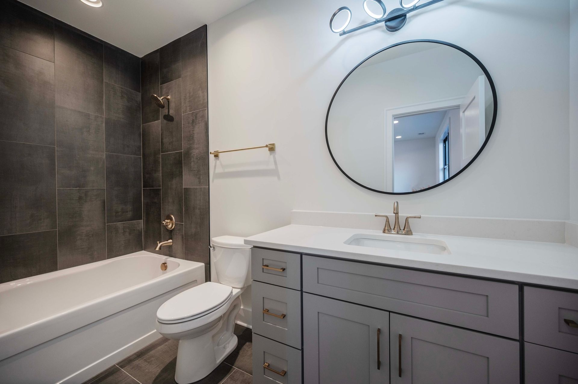 Modern bathroom with gray vanity, round mirror, and dark tiled bath.