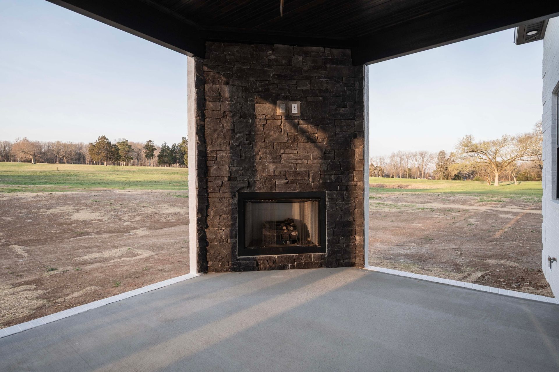 Outdoor corner with fireplace, facing a grassy field under a clear sky.