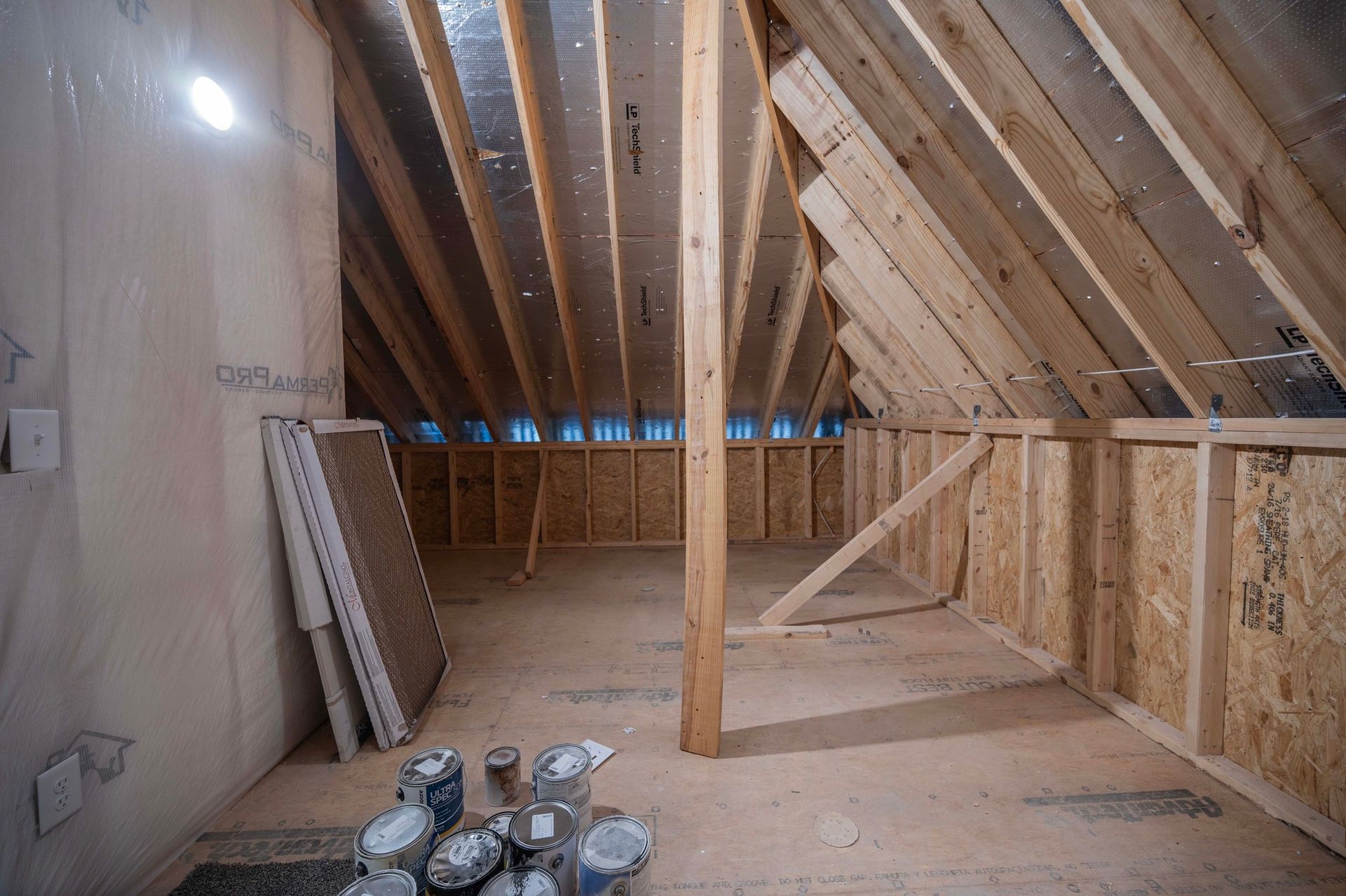 Interior of unfinished attic with exposed wooden rafters and framing, materials, and paint cans.