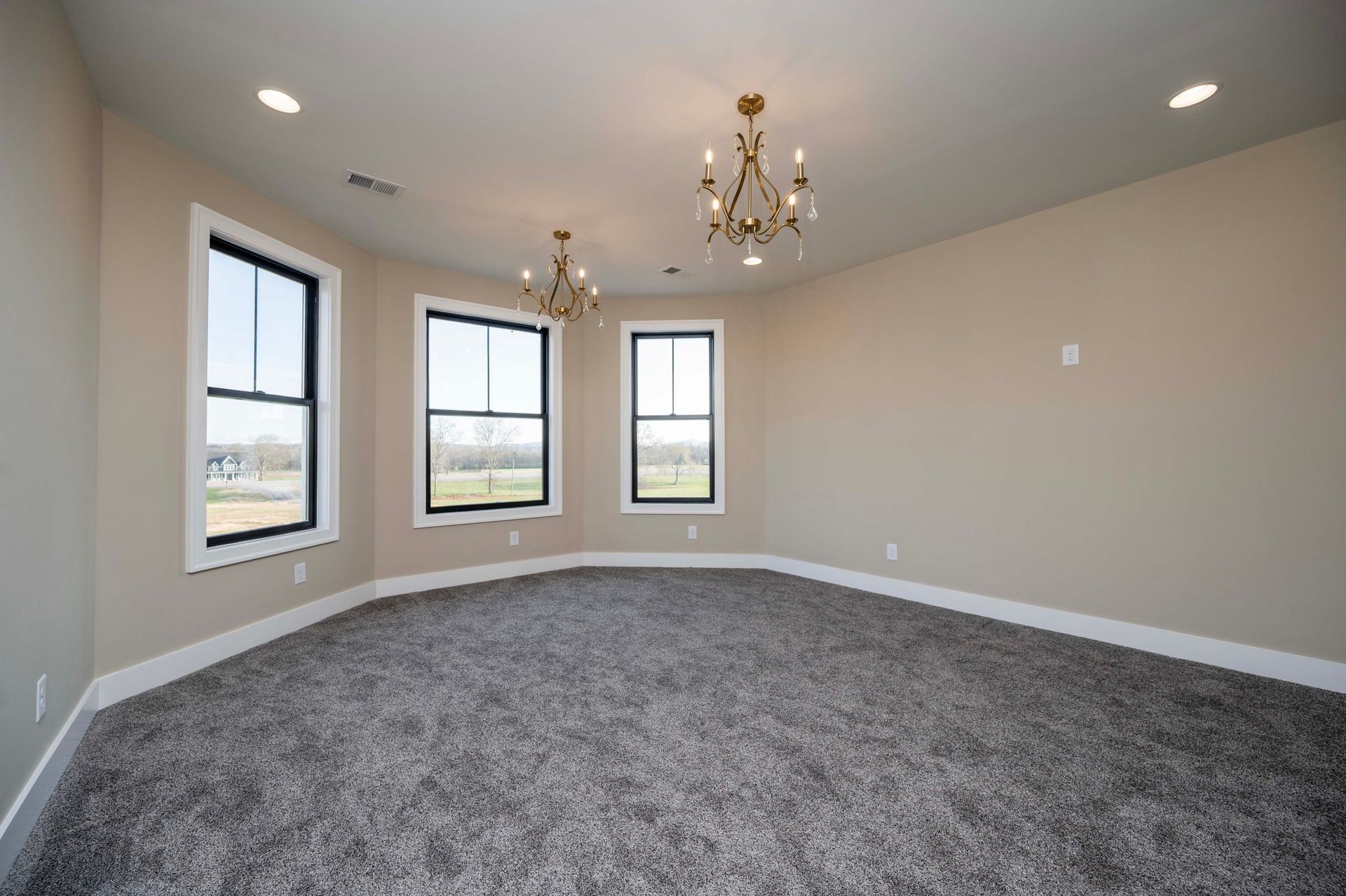 Empty room with three windows, two chandeliers, and gray carpet. Beige walls and white trim.