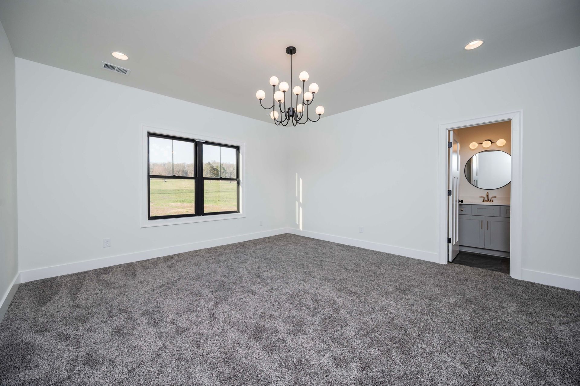 Empty bedroom with gray carpet, black-framed window, and a chandelier. Bathroom visible through an open door.