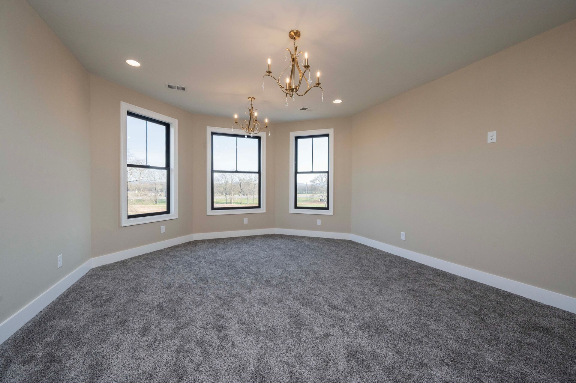 Empty room with gray carpet, beige walls, three windows, two chandeliers.