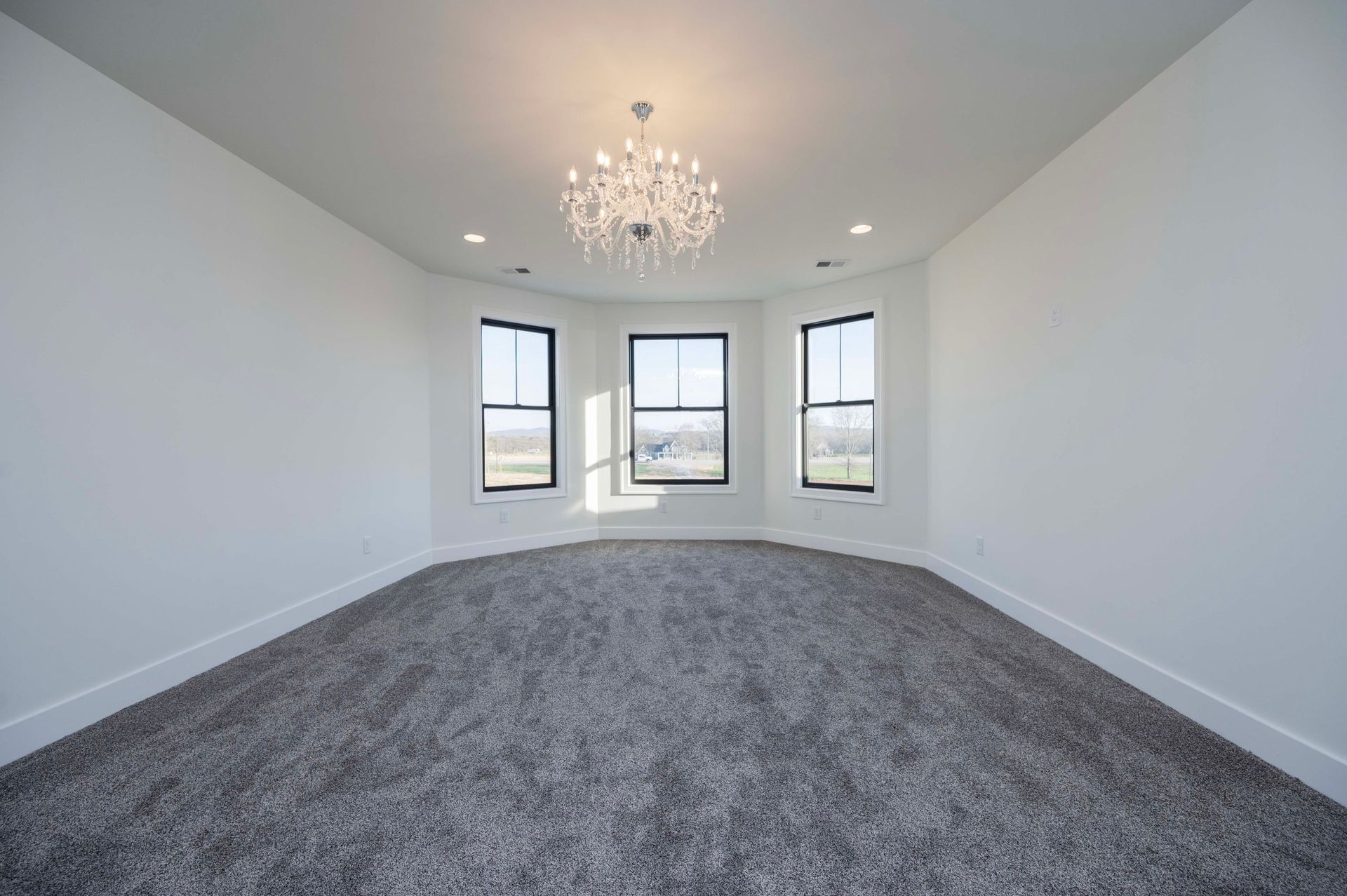 Empty room with grey carpet, white walls, and a bay window. Crystal chandelier hangs from the ceiling.